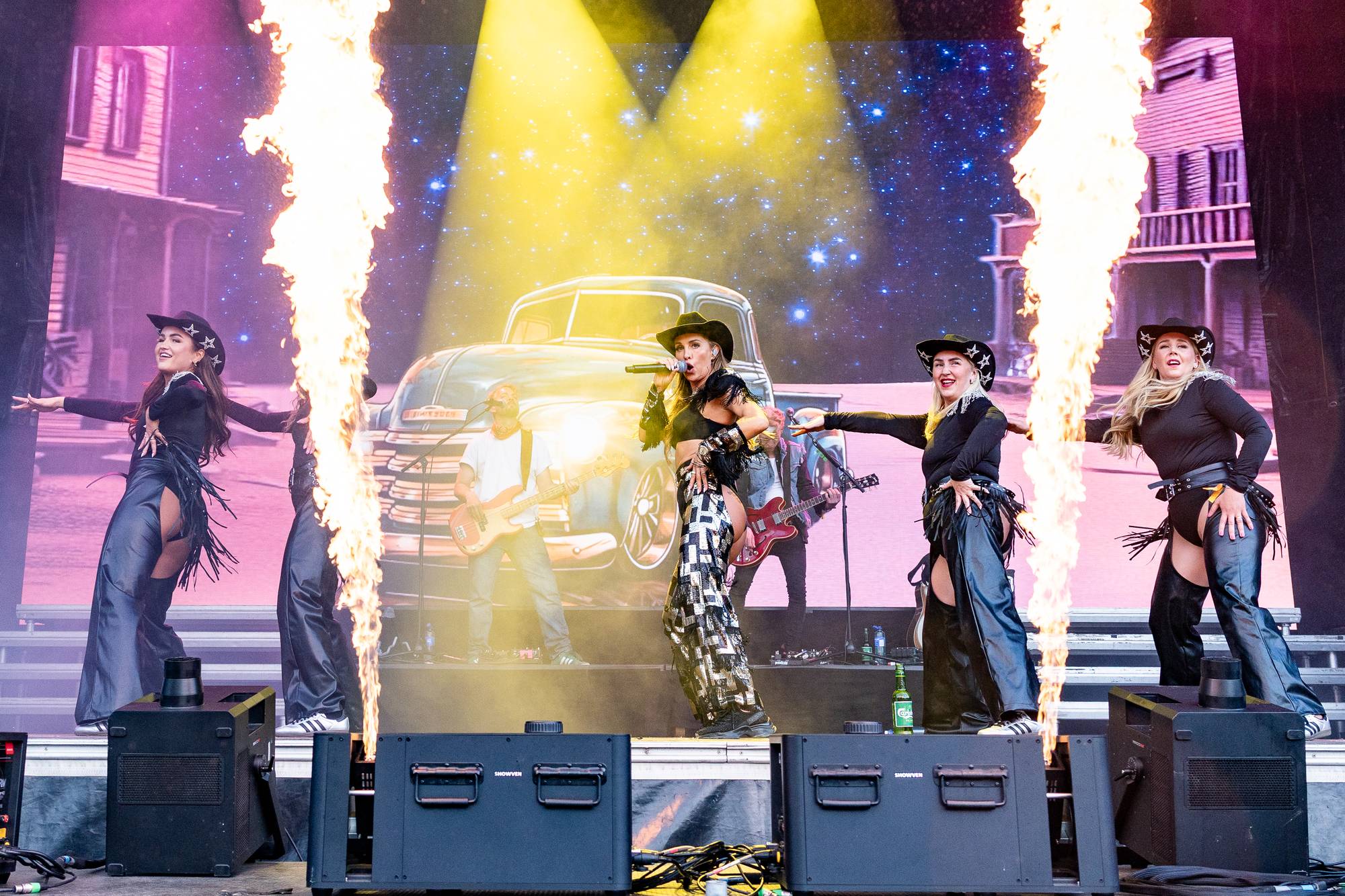 Scenen er dekorert som en westernby med flammekastere. En kvinne, kledd i cowboyhatt og matchende antrekk, synger i et headset mens hun holder en mikrofon. Rundt henne danser tre andre kvinner. En mann spiller gitar bak henne. (Bildebeskrivelsen er laget av en KI-tjeneste)