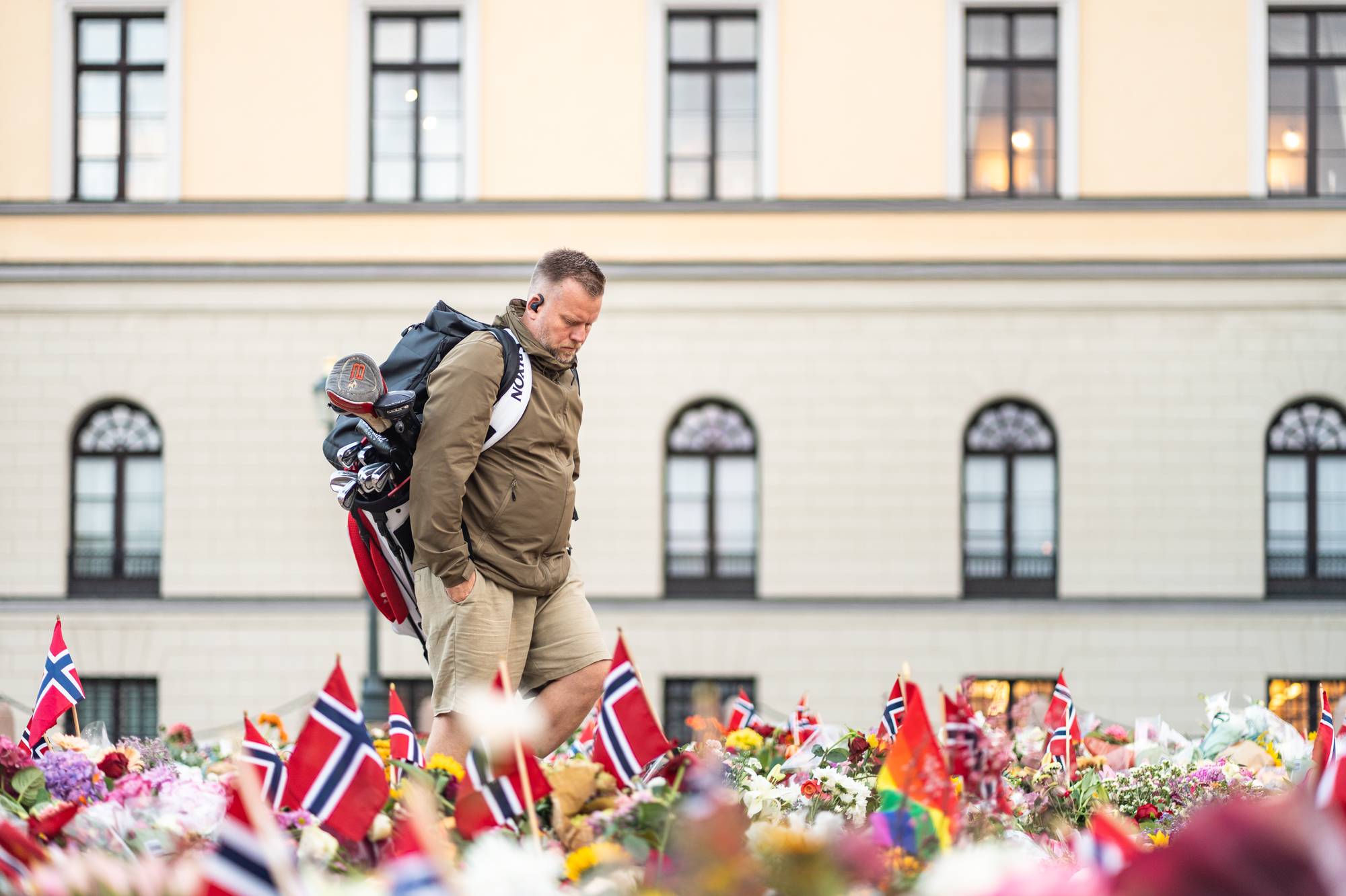 En mann med golfbag går forbi et hav av blomster og norske flagg foran en bygning. (Bildebeskrivelsen er laget av en KI-tjeneste)