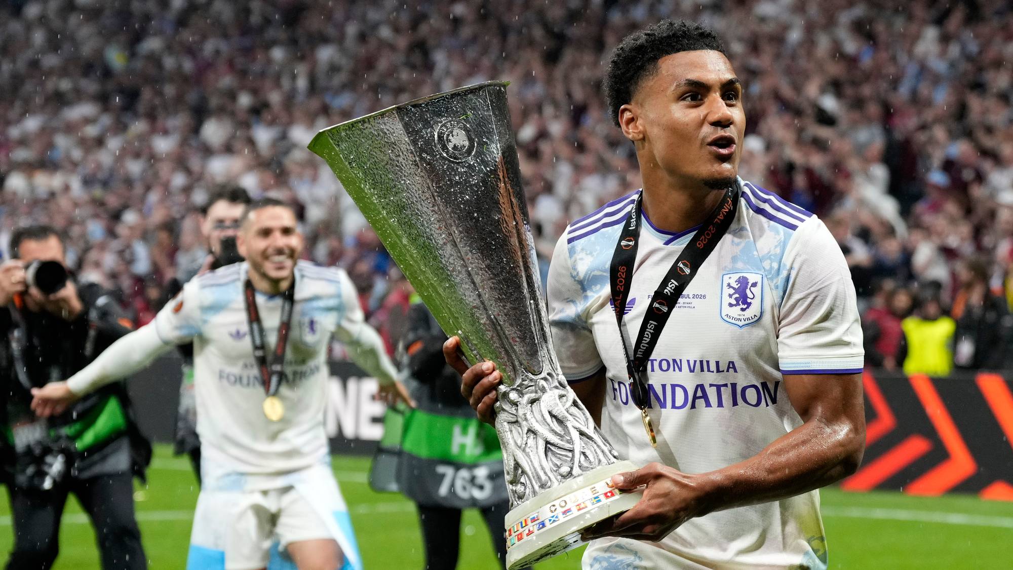 Fotballspiller holder Europa League-trofeet etter seier på stadion med jublende lagkamerater bak. (Bildebeskrivelsen er laget av en KI-tjeneste)