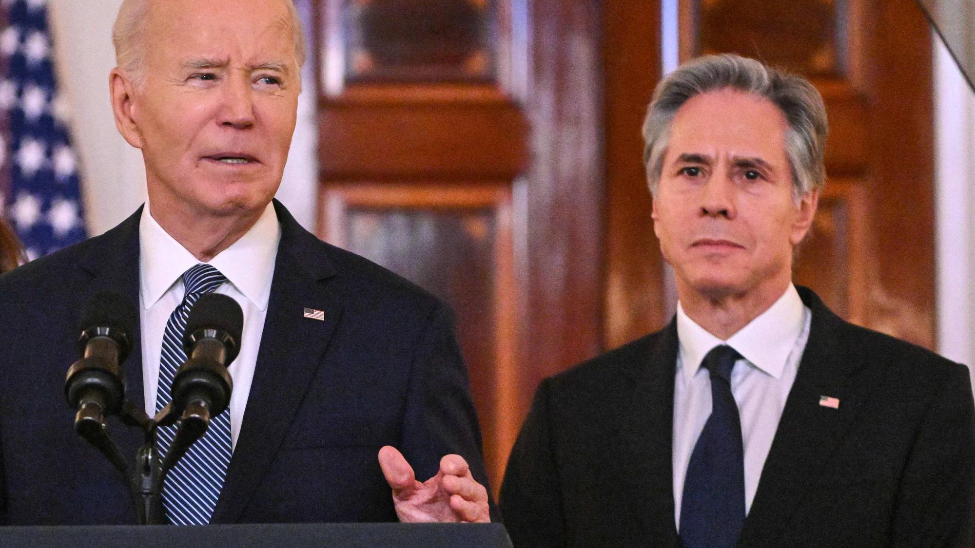 USAs president Joe Biden taler ved et podium, mens utenriksminister Antony Blinken står ved siden av. Begge er kledd i mørke dresser og slips. Bak dem er en amerikansk flagg. (Bildebeskrivelsen er laget av en KI-tjeneste)