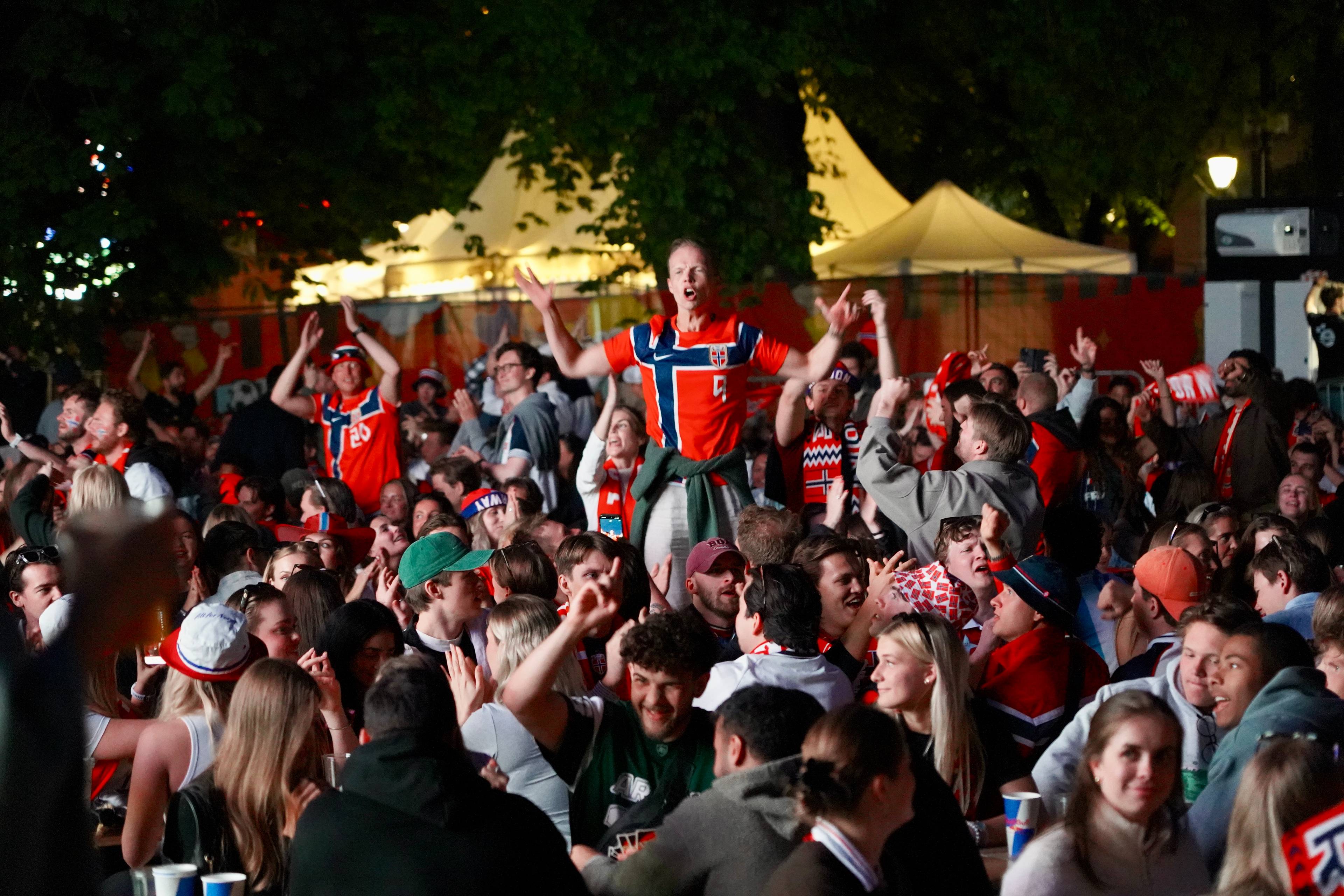 En stor folkemengde med supportere i norske farger ser på en skjerm utendørs. En mann står på skuldrene til noen andre og jubler. Mange har på seg supporterutstyr som vimpler og bannere med norske flagg. Omgivelsene er mørke, og det er telt og lys i bakgrunnen.