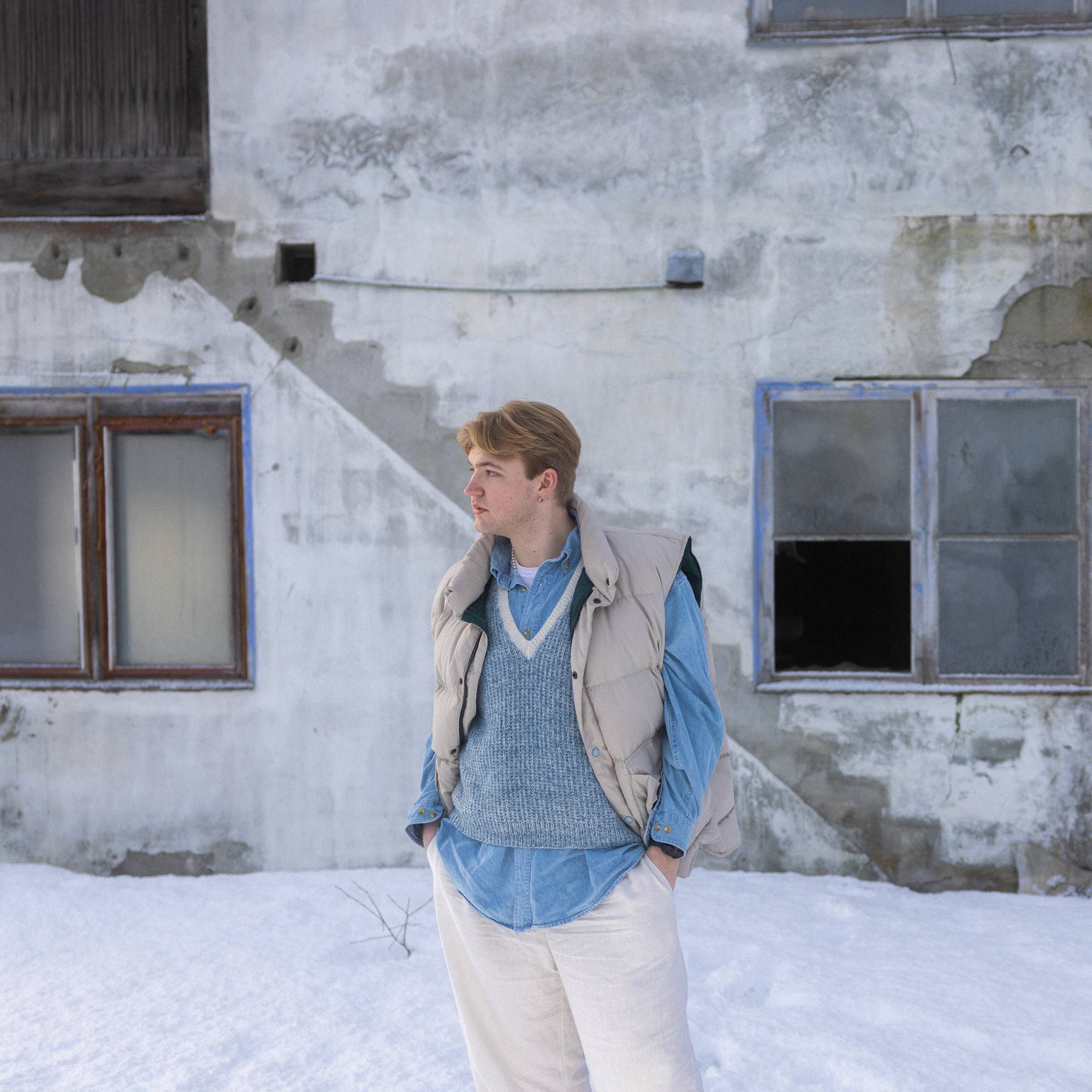 Viktor står foran en forlatt bygning med de samme fargene på antrekket sitt, ser ut til siden tenkende med hendene i lommene. Det er kald vinter ute, snø på bakken.