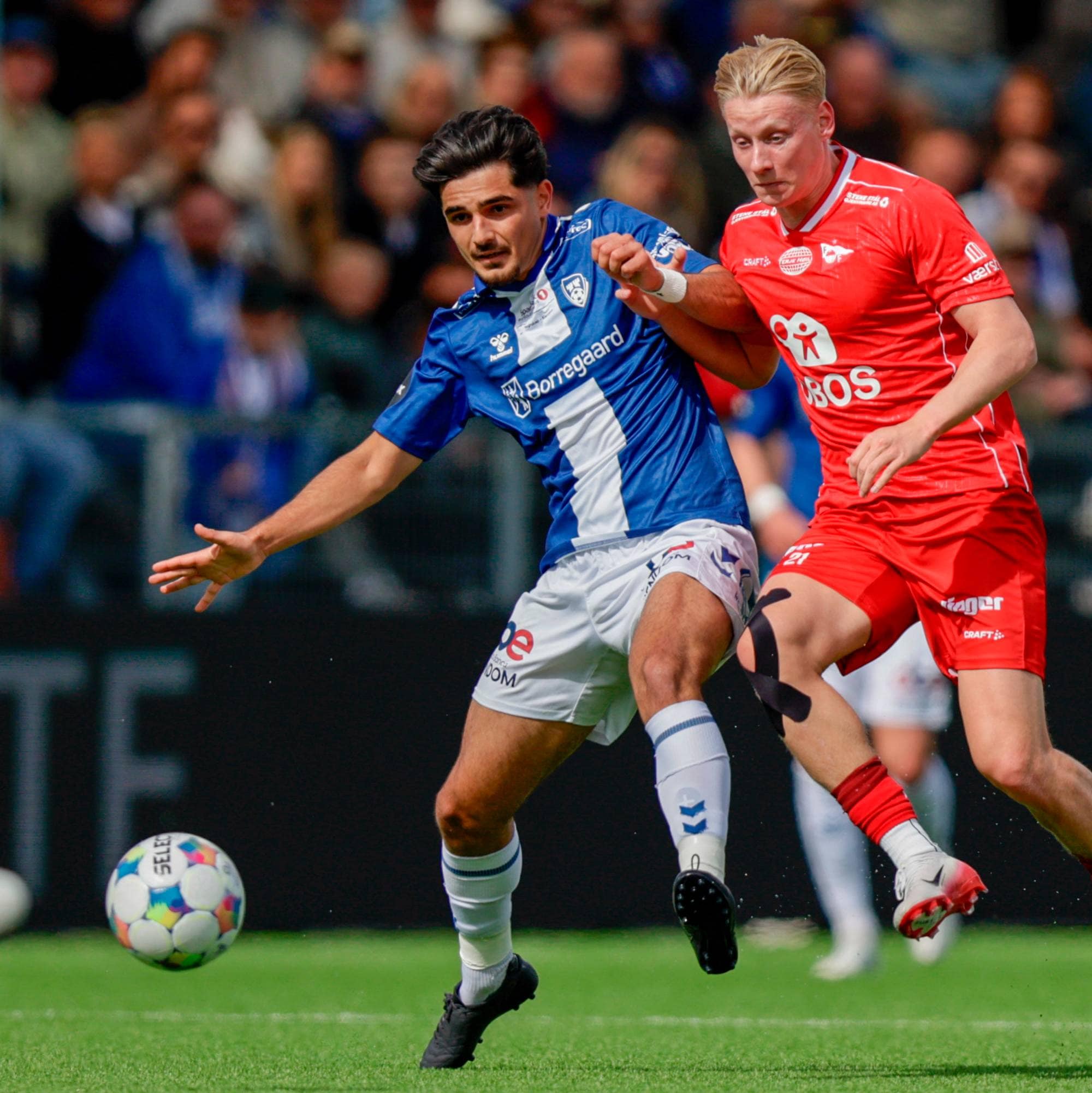 To fotballspillere kjemper om ballen på en grønn gressbane. Den ene spilleren har blå drakt med hvite detaljer, den andre har rød drakt. En fotball ligger i forgrunnen. Tilskuerne er uskarpe i bakgrunnen. (Bildebeskrivelsen er laget av en KI-tjeneste)