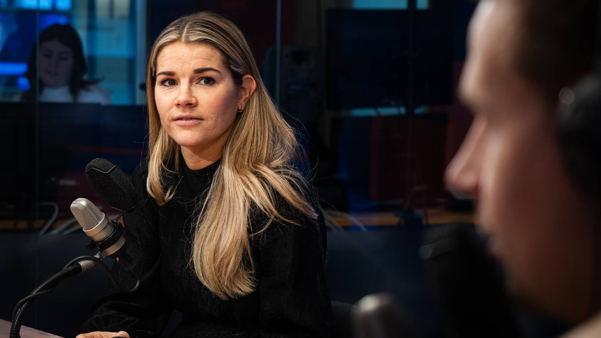 En kvinne sitter foran en mikrofon i et studio. Bak henne er en glassvegg med speilbilde av en annen person. En person med headset er uskarpt i forgrunnen til høyre. (Bildebeskrivelsen er laget av en KI-tjeneste)