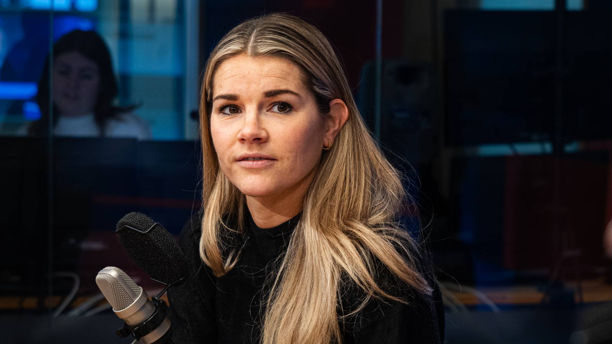 En kvinne sitter foran en mikrofon i et studio. Bak henne er en glassvegg med speilbilde av en annen person. En person med headset er uskarpt i forgrunnen til høyre. (Bildebeskrivelsen er laget av en KI-tjeneste)