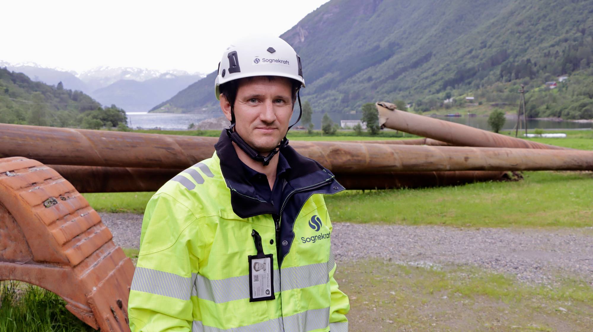 Mann i verneutstyr foran store rør ved et vann. Bakgrunnen viser fjell og natur. (Bildebeskrivelsen er laget av en KI-tjeneste)