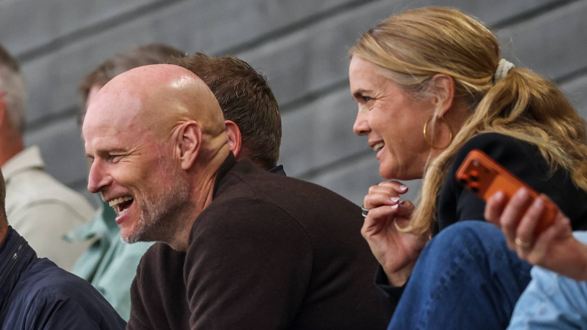 Tilskuere sitter på en tribune under en idrettsbegivenhet. Flere menn og en kvinne er synlige i forgrunnen. De ser ut til å følge med på noe utenfor bildet. Bakgrunnen viser en grå tre-lignende overflate. Tilskuere sitter tett i tett på tribunen, noe som indikerer en begivenhet med mange tilskuere. (Bildebeskrivelsen er laget av en KI-tjeneste)