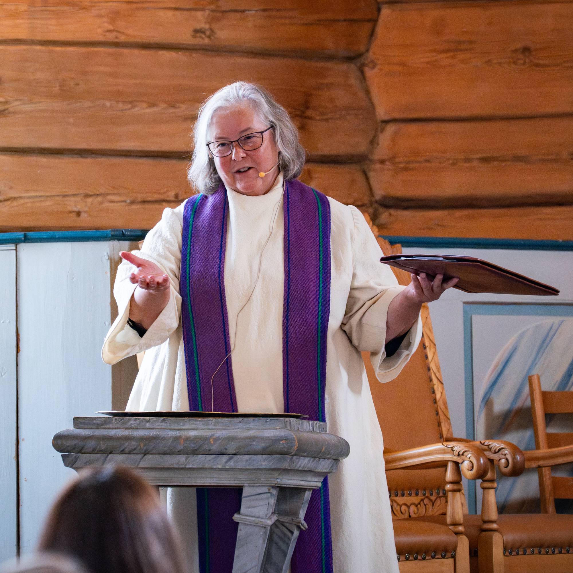 Prest i hvit kappe og lilla stola taler fra en talerstol i en kirke med tømmervegger. (Bildebeskrivelsen er laget av en KI-tjeneste)