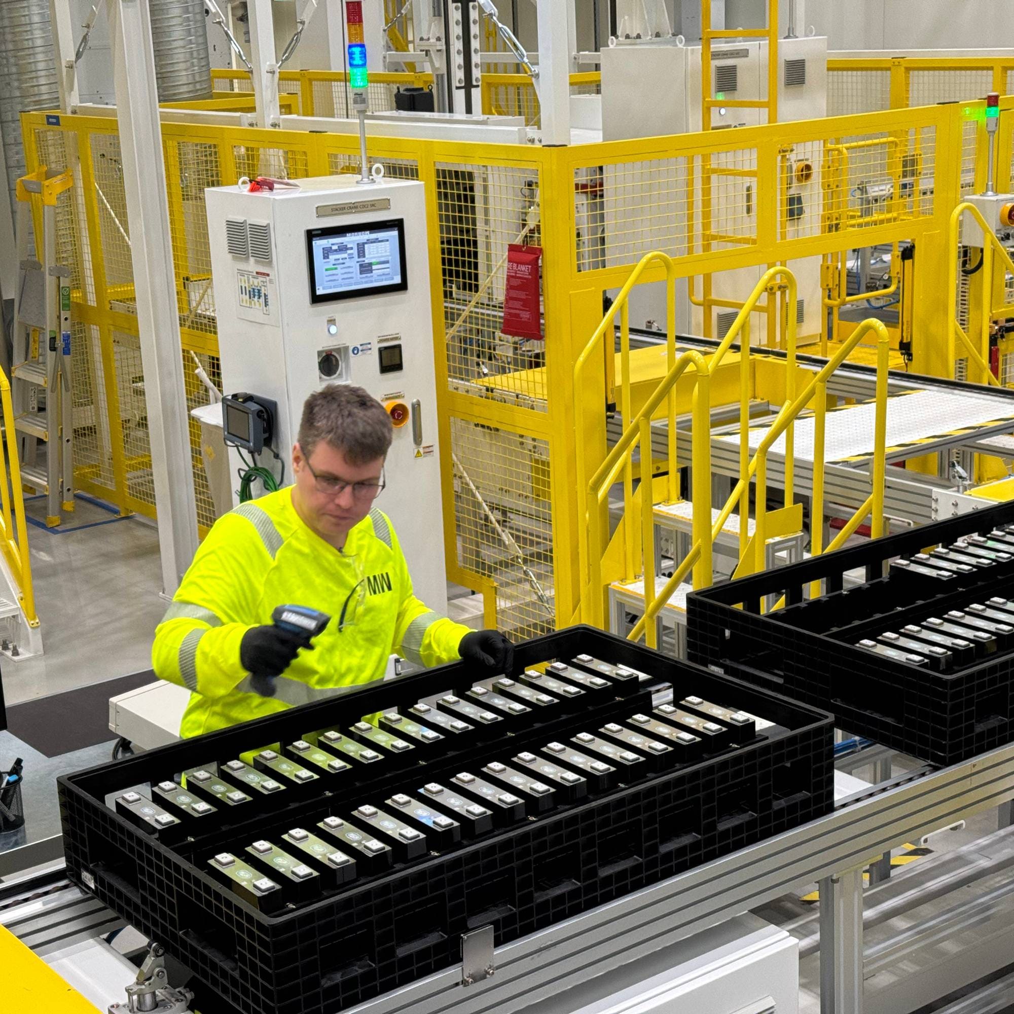 Ansatt i Morrow sorterer ferdige battericeller på samlebånd. Har gul vest, og fabrikkutstyr i bakgrunnen. 