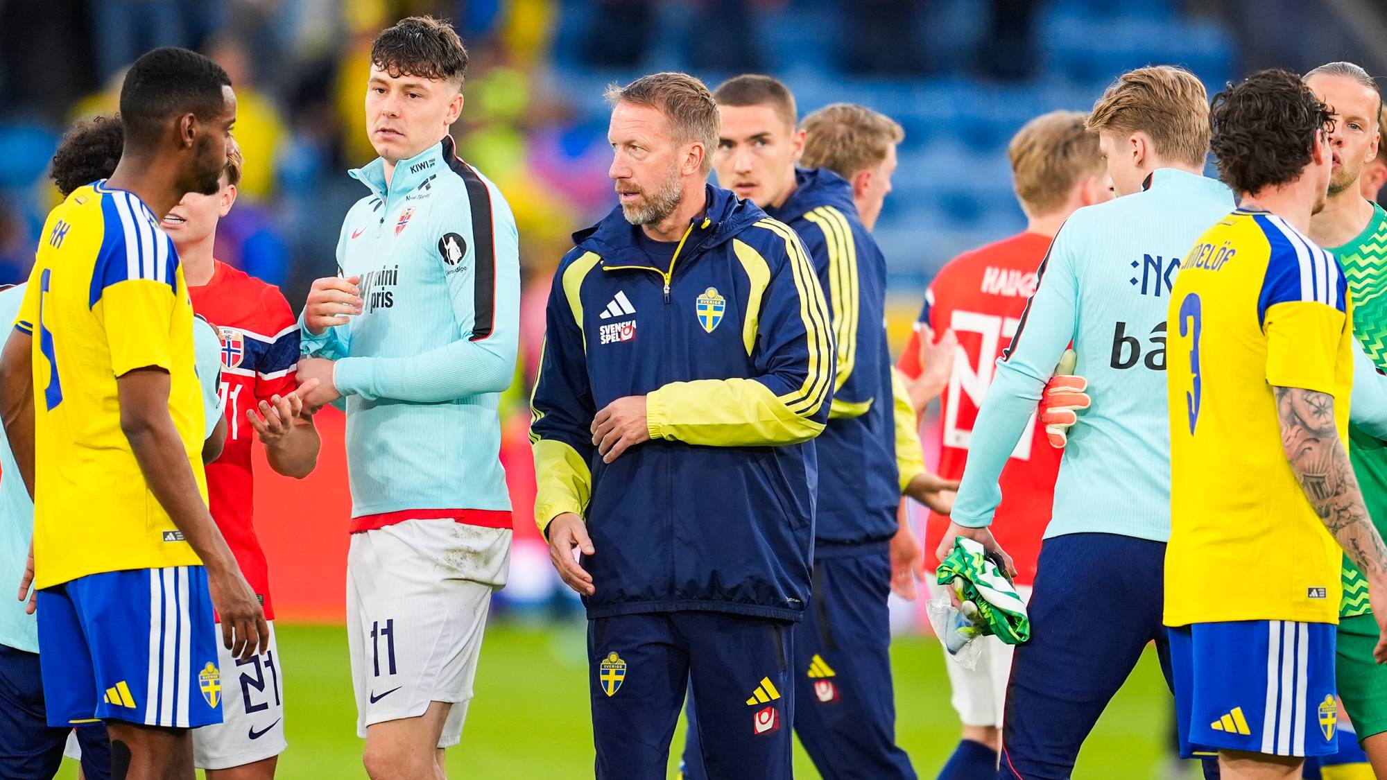 En gruppe fotballspillere og trenere samlet på en bane. En av dem, i mørkeblå jakke med gule detaljer, er trolig trener. Flere spillere er iført gule og blå drakter, mens andre har på seg lyseblå og røde drakter. Bildet ser ut til å være tatt på en stadion. (Bildebeskrivelsen er laget av en KI-tjeneste)