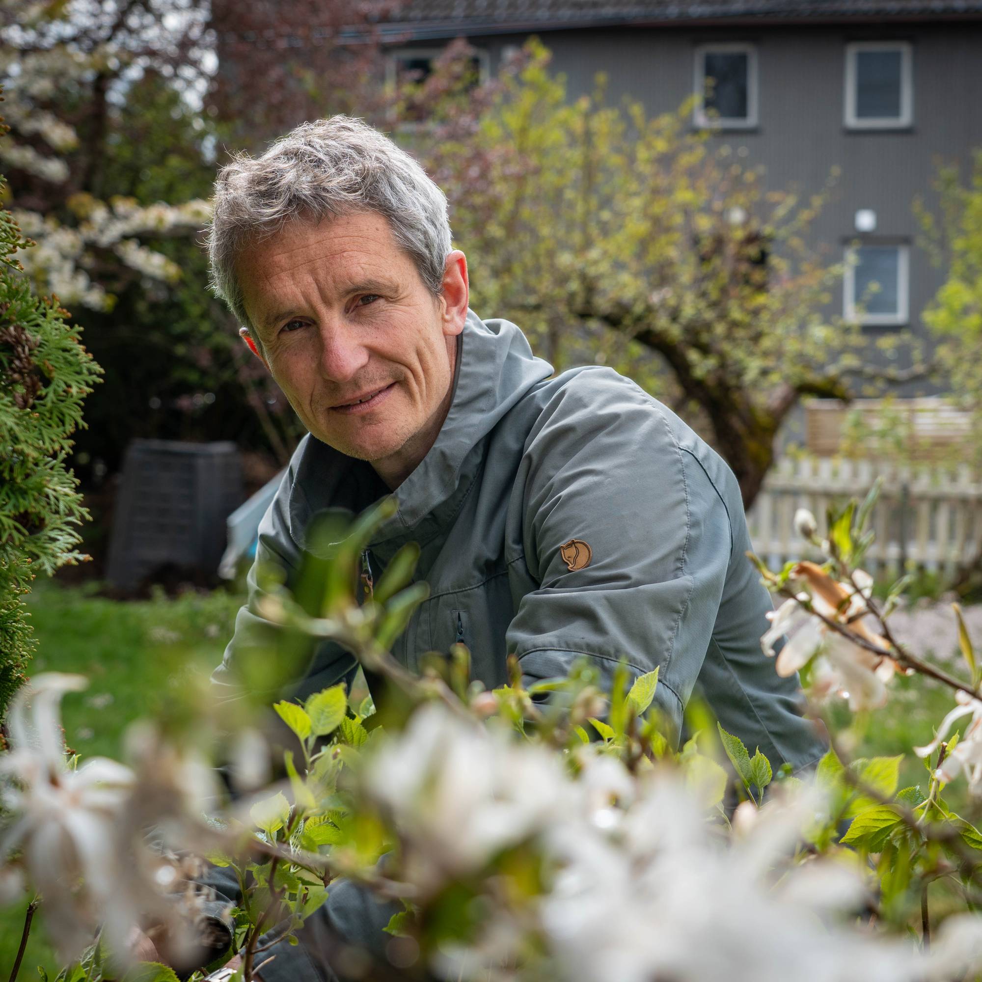 Middelaldrende mann i grå jakke poserer i en hage med busker og blomster.
