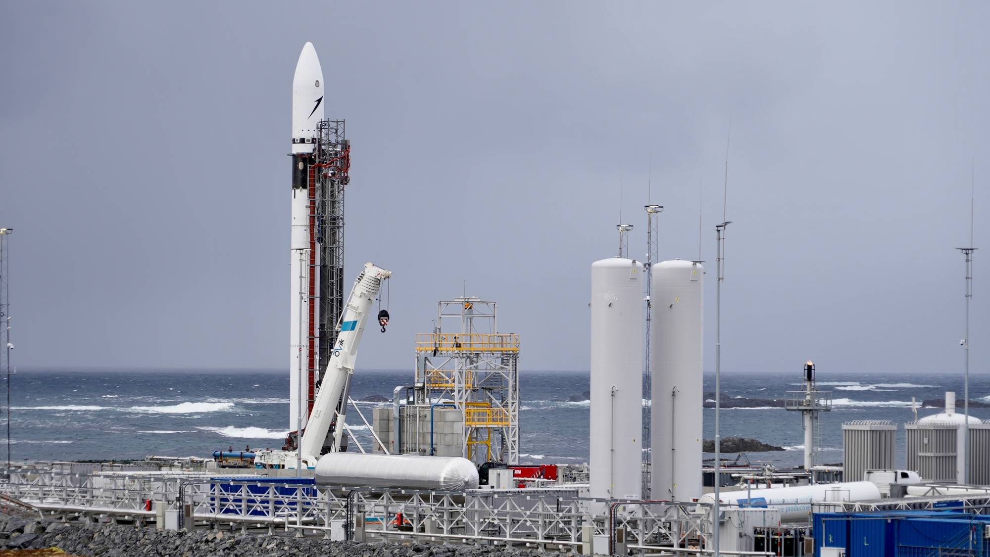 En rakett står klar på en oppskytningsplattform ved havet. En kran er plassert ved siden av raketten. Flere store tanker er synlige på plattformen. En person er synlig på en av plattformene. (Bildebeskrivelsen er laget av en KI-tjeneste)
