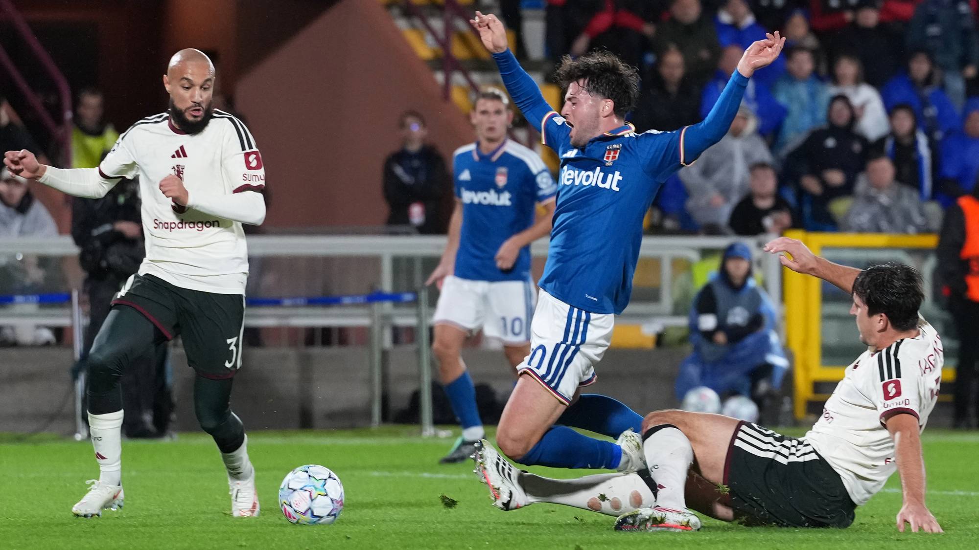 Fotballspillere i duell på banen under en kamp. En spiller i blått faller i gresset. (Bildebeskrivelsen er laget av en KI-tjeneste)