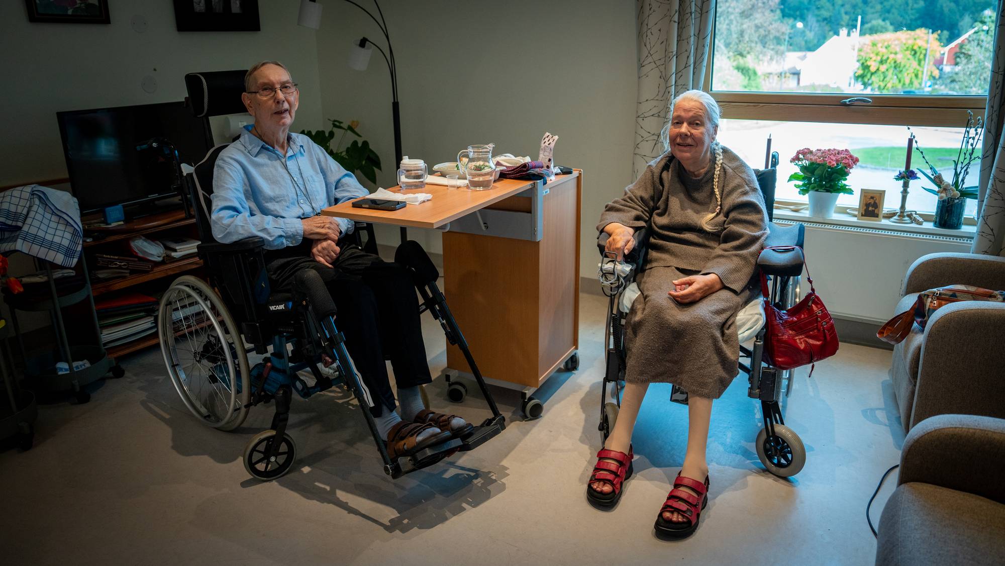 To eldre mennesker i rullestol sitter sammen i et rom. (Bildebeskrivelsen er laget av en KI-tjeneste)