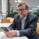 Håvard Axel Kator / NRK Gjøsæter har på seg briller og tweedjakke og sitter og ser på mobilen. Han sitter ved et hvitt bord med gule stoler rundt. I bakgrunnen er det planter og bord.