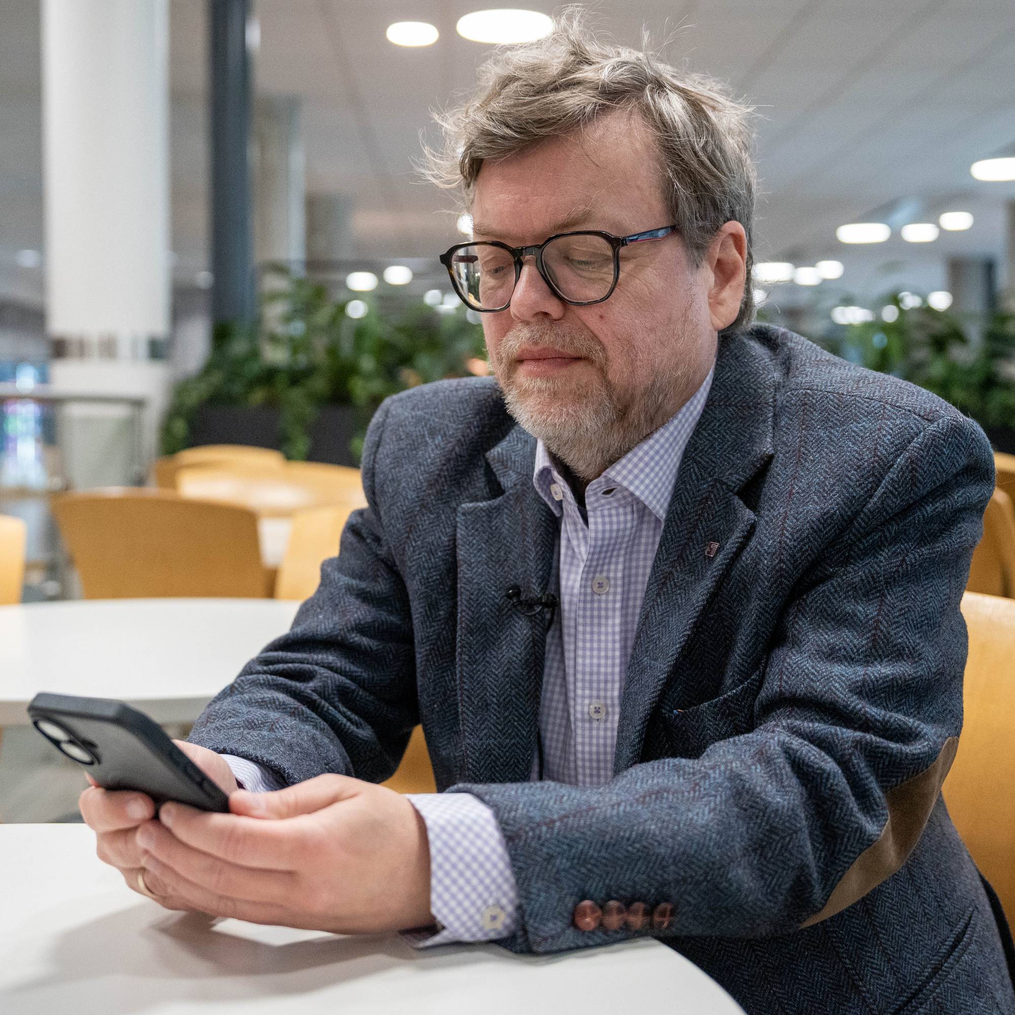 Gjøsæter har på seg briller og tweedjakke og sitter og ser på mobilen. Han sitter ved et hvitt bord med gule stoler rundt. I bakgrunnen er det planter og bord. 