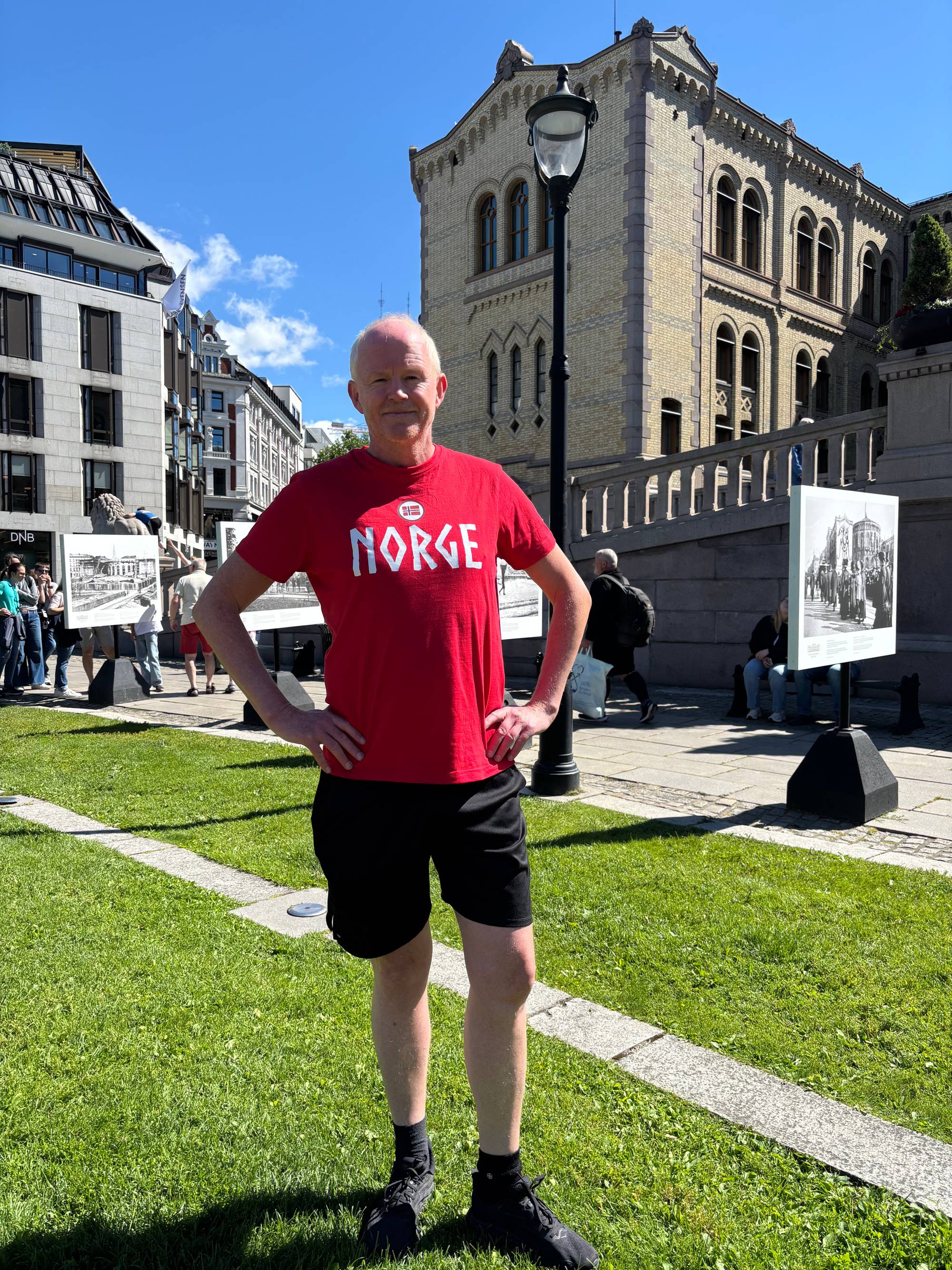 En middelaldrende mann i rød t-skjorte med Norge skrevet med store, hvite bokstaver på brystet står på en grønn gressplen foran Stortinget, smilende med hendene på hoftene og ser i kameraet.