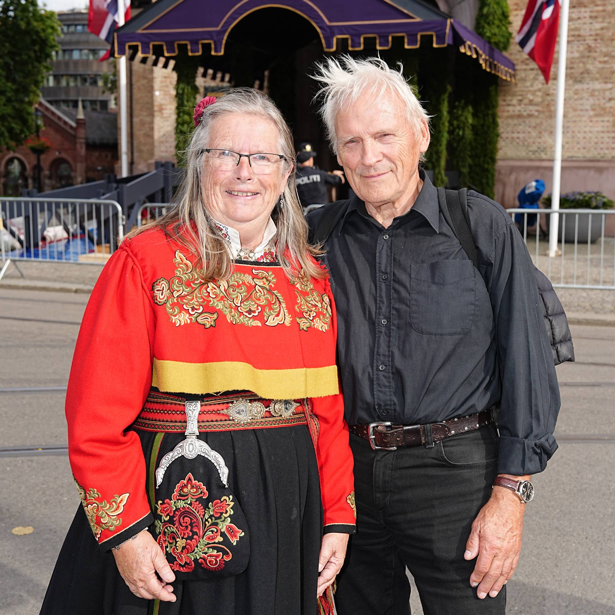 Kvinne i bunad og mann i svarte klær står foran Oslo domkirke. Norske flagg i bakgrunnen. (Bildebeskrivelsen er laget av en KI-tjeneste)