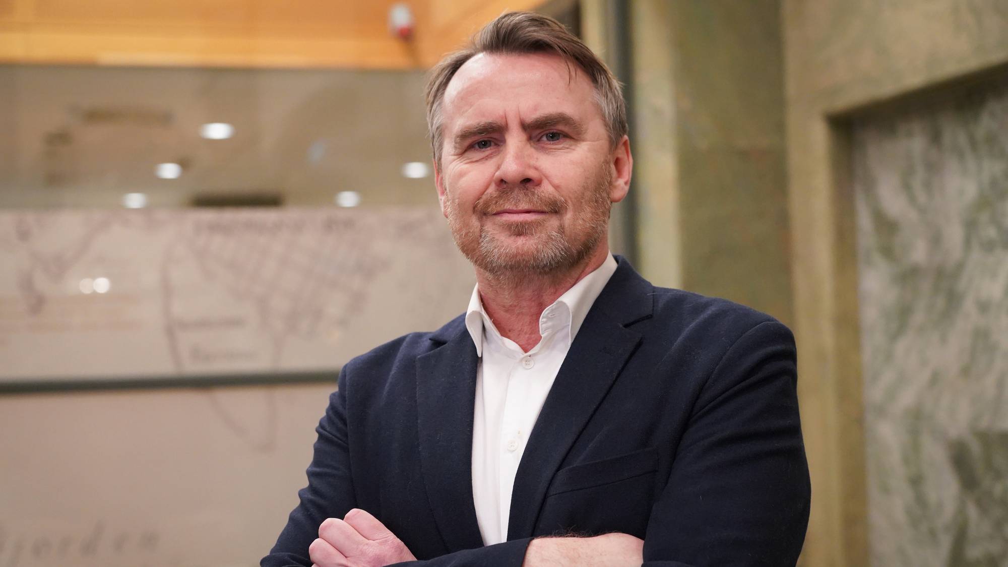 En mann står med armene krysset foran brystet, iført blazer og skjorte. Bakgrunnen består av en mørk vegg med tekst og grafikk. Det er innendørs belysning, som gir et lyst miljø. Mannen har et alvorlig uttrykk.