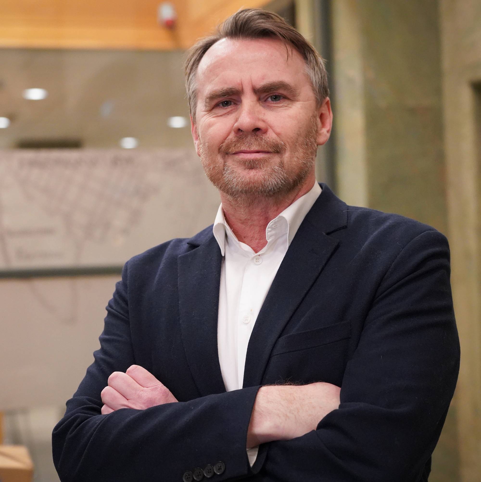 En mann står med armene krysset foran brystet, iført blazer og skjorte. Bakgrunnen består av en mørk vegg med tekst og grafikk. Det er innendørs belysning, som gir et lyst miljø. Mannen har et alvorlig uttrykk.