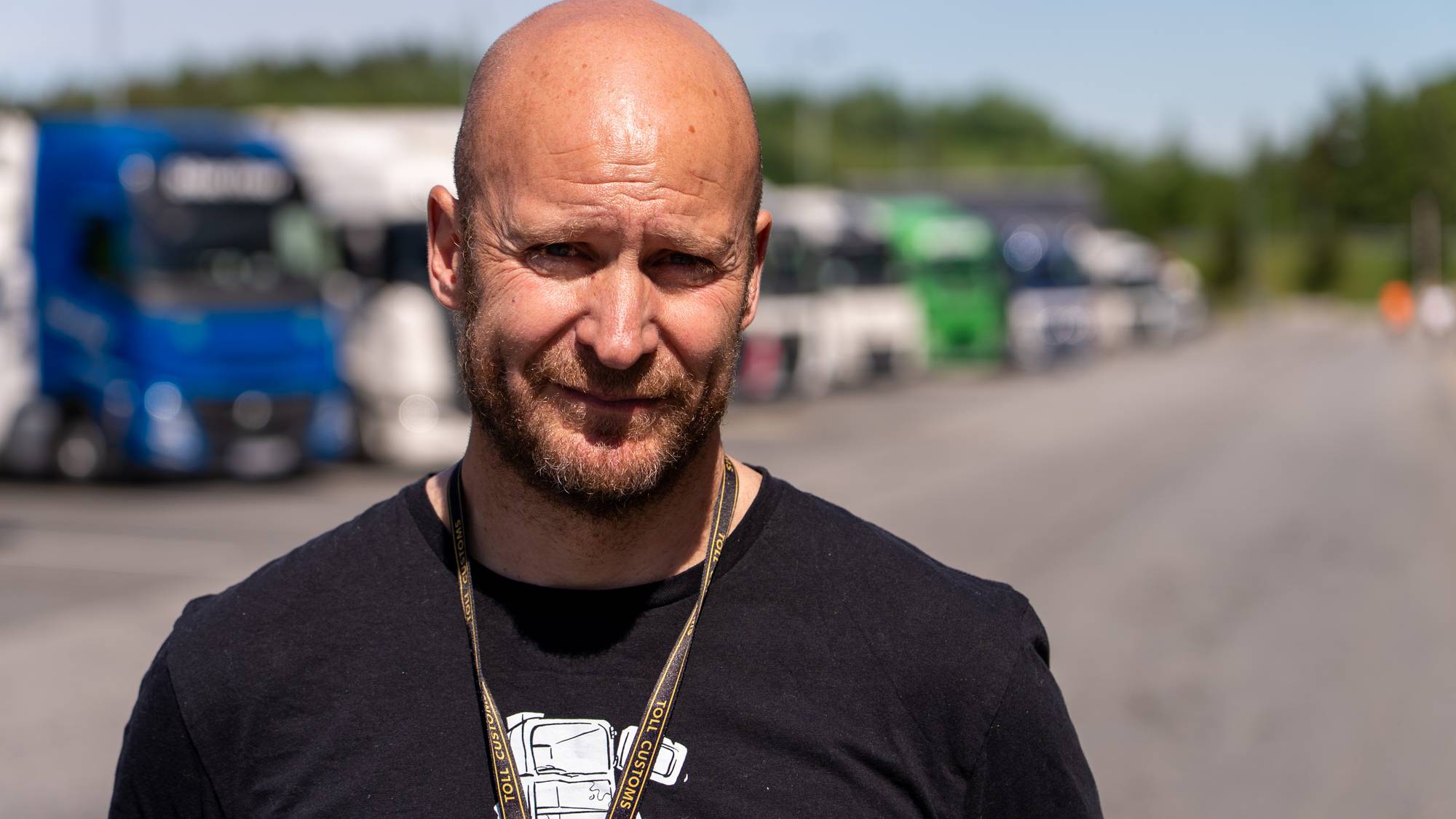 Bald man with beard wearing black t-shirt.
He wears a lanyard with "Toll Customs" written on it.
In the background, there are parked trucks.
The photo is taken outdoors on a sunny day. (Bildebeskrivelsen er laget av en KI-tjeneste)