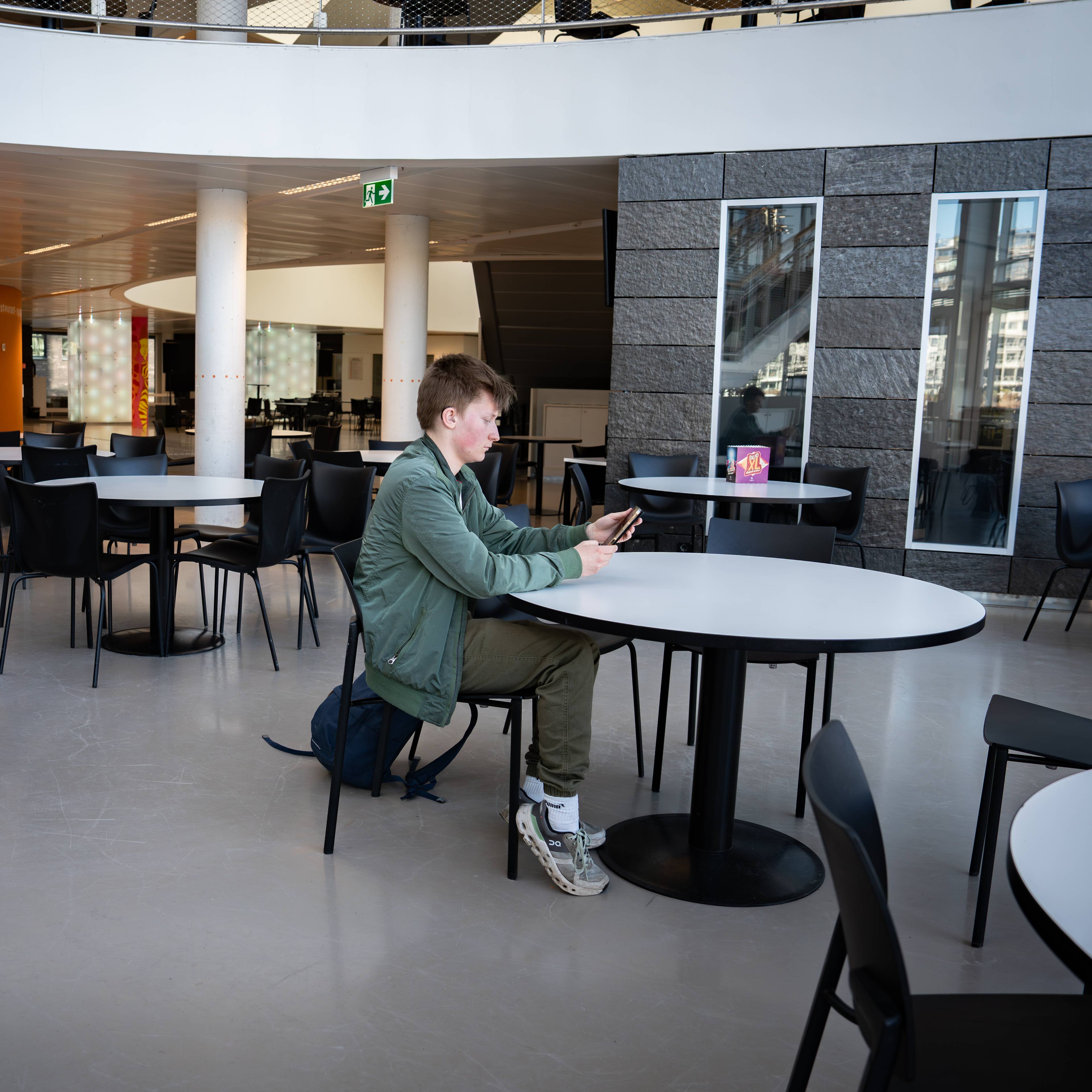 Christian sitter alene ved et rundt bord på en kaf. Han ser ned på mobiltelefonen sin. I bakgrunnen er det flere bord og stoler, samt en heis.