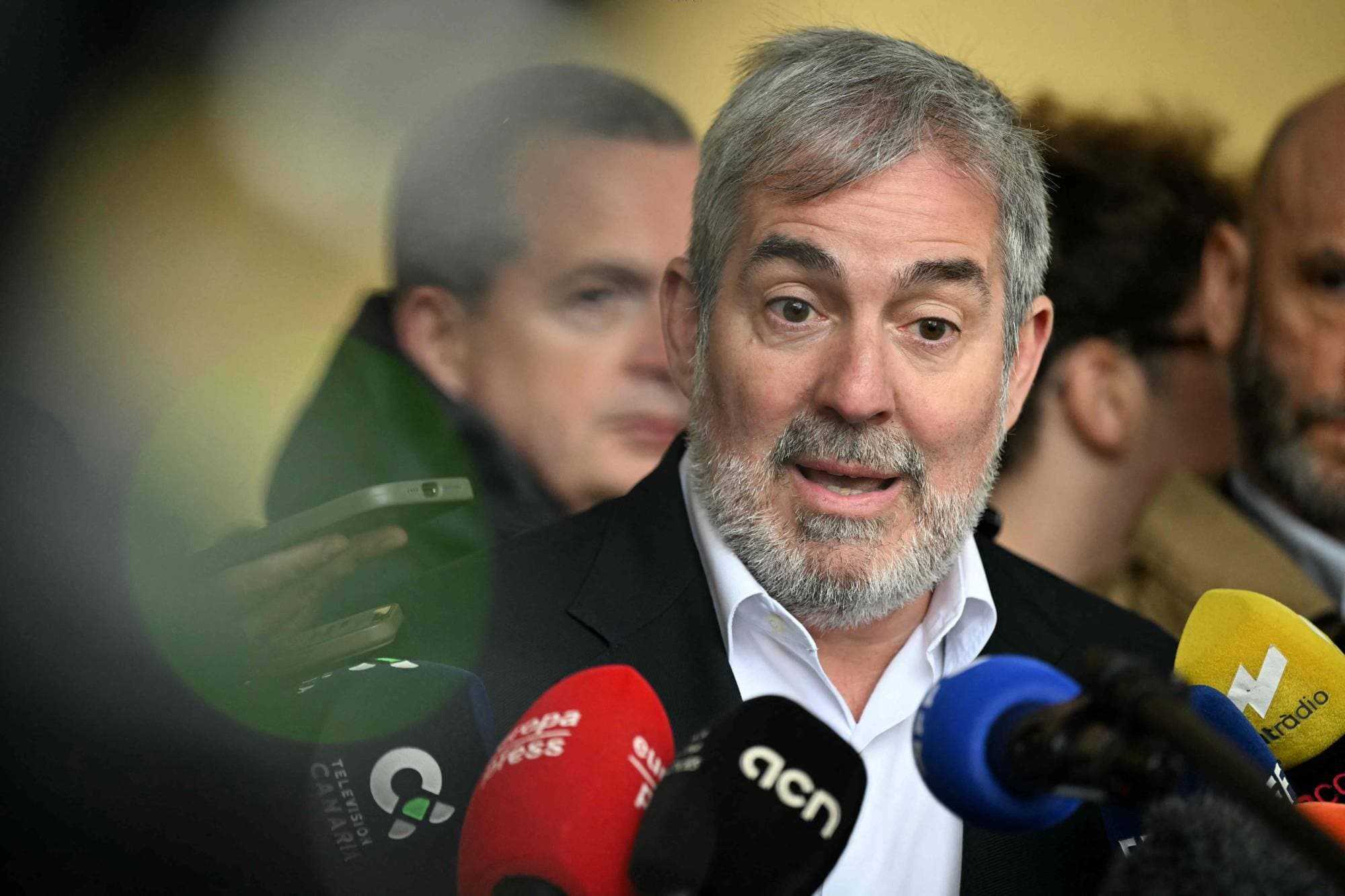 En mann med grått skjegg snakker foran flere mikrofoner. Bak ham sees utydelige personer. En hånd holder en mobiltelefon. (Bildebeskrivelsen er laget av en KI-tjeneste)