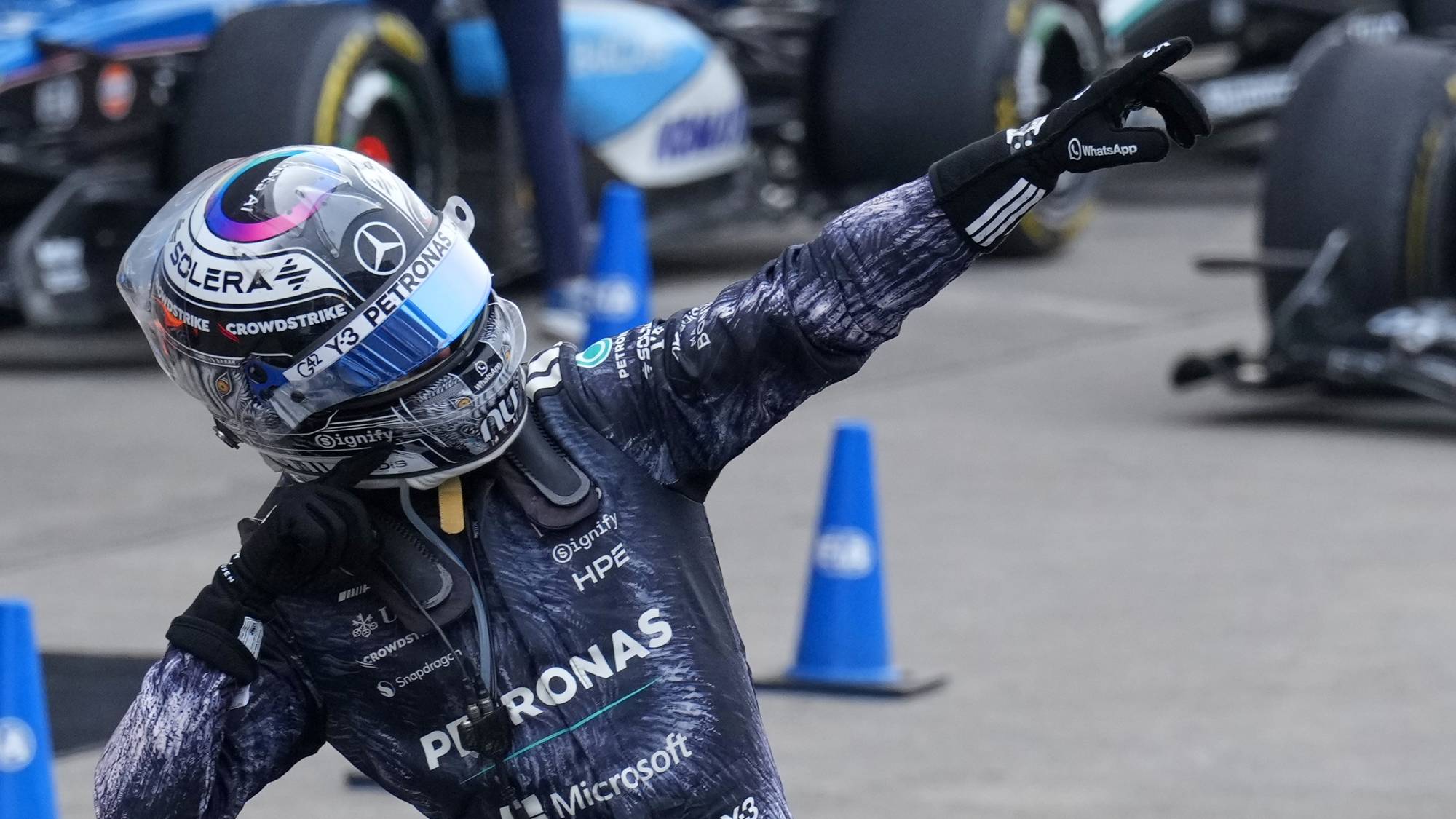 En racerfører i hjelm og drakt peker med høyre hånd. Bak ham står en Formel 1-bil. Det er flere kjegler og en annen bil i bakgrunnen. Deler av en annen bil er synlig nederst i bildet. (Bildebeskrivelsen er laget av en KI-tjeneste)