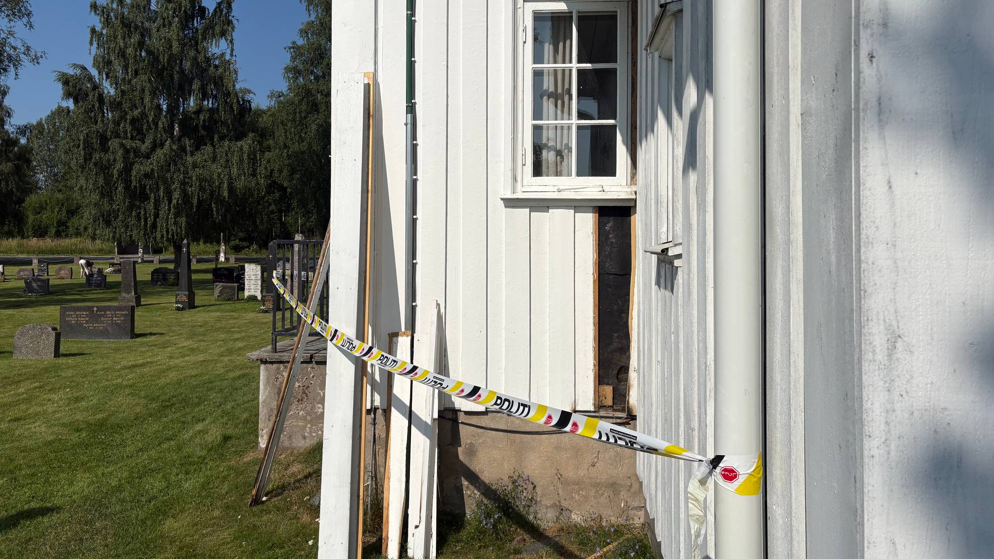 Politibånd er sperret opp foran et åpnet veggpanel på en hvit trebygning ved en kirkegård. (Bildebeskrivelsen er laget av en KI-tjeneste)