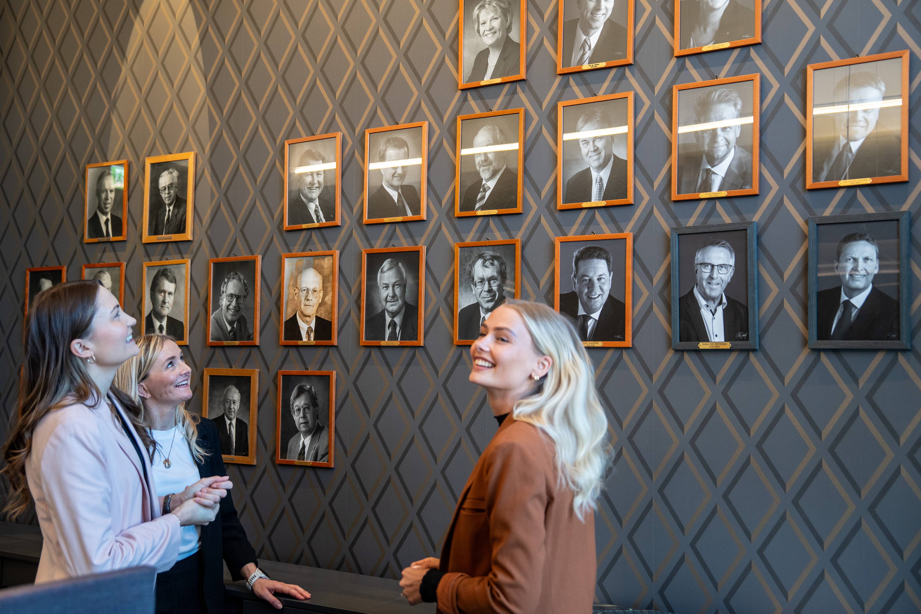 Tre kvinner ser på en vegg med portrettbilder i rammer. Bildene viser eldre menn. En av kvinnene er i forgrunnen og smiler. (Bildebeskrivelsen er laget av en KI-tjeneste)