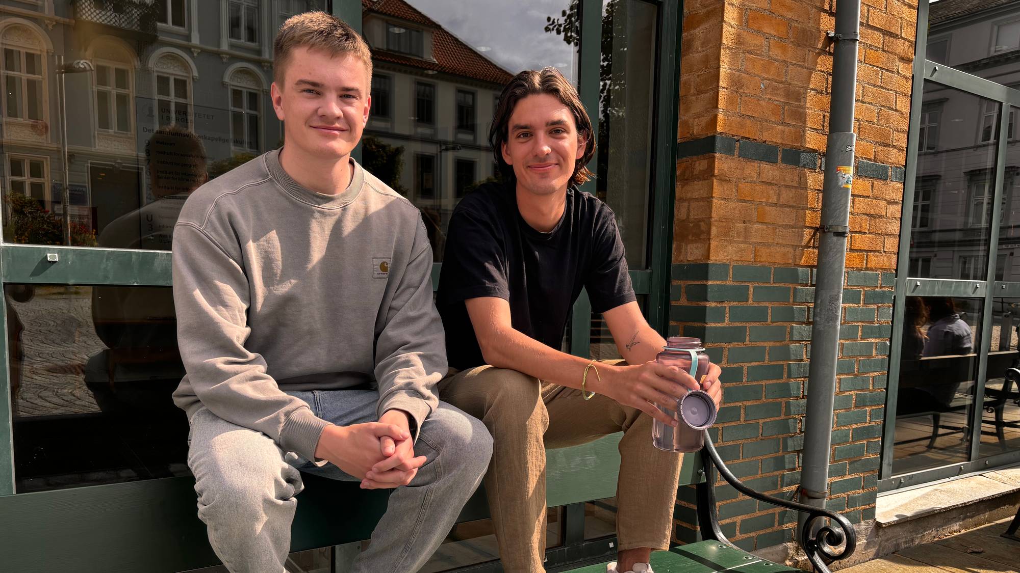 To unge menn sitter på en benk utenfor en bygning med mursteinsfasade. Den ene mannen har på seg en grå genser og jeans, mens den andre har på seg en svart t-skjorte og lysebrune bukser. Begge ser mot kameraet. Bak dem er det et stort vindu og en murvegg. (Bildebeskrivelsen er laget av en KI-tjeneste)
