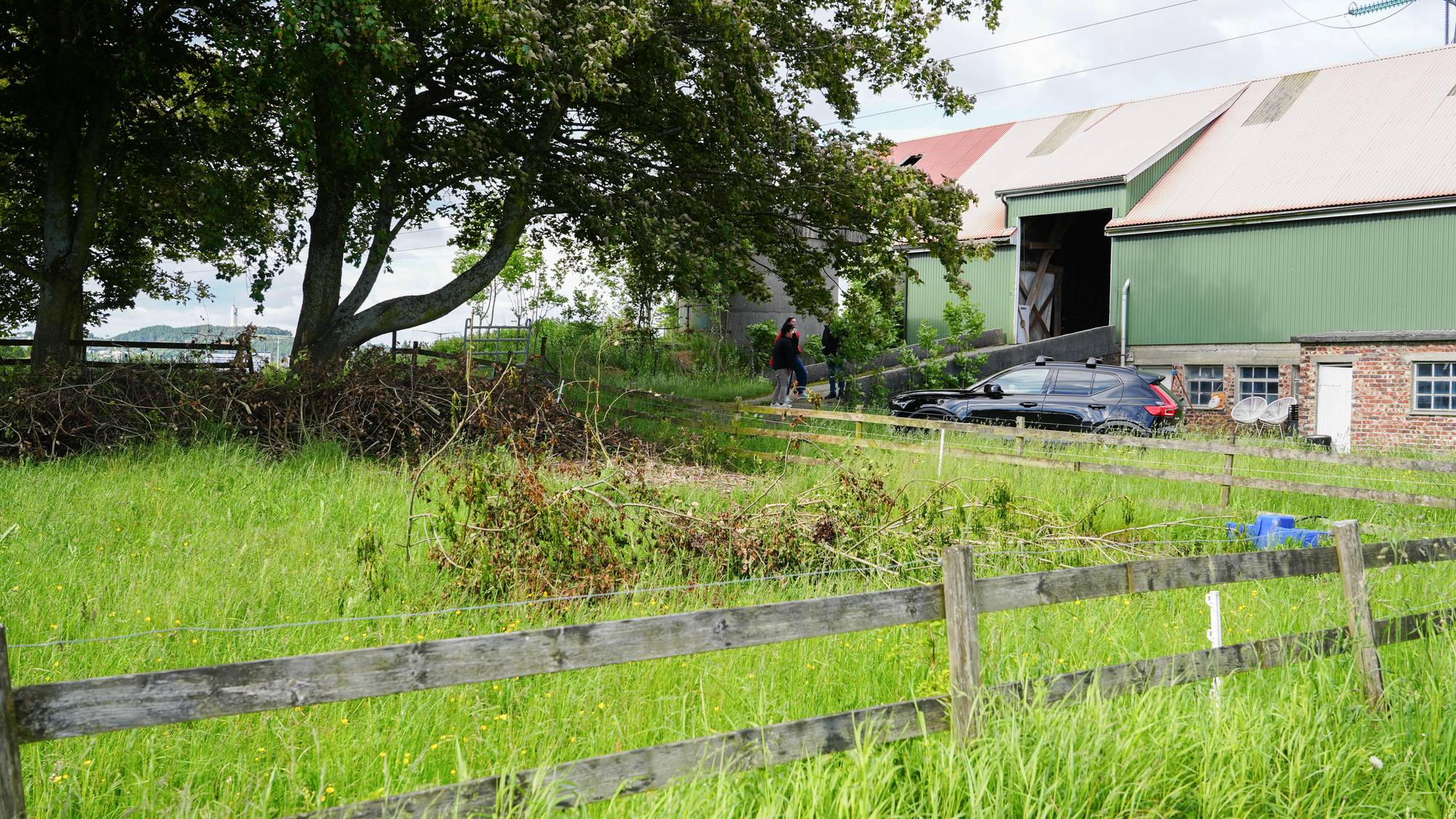 Tre personer står ved et gjerde foran en gård. En svart bil er parkert ved siden av en grønn bygning. Området rundt er preget av frodig gress og et tre. Været er overskyet. (Bildebeskrivelsen er laget av en KI-tjeneste)
