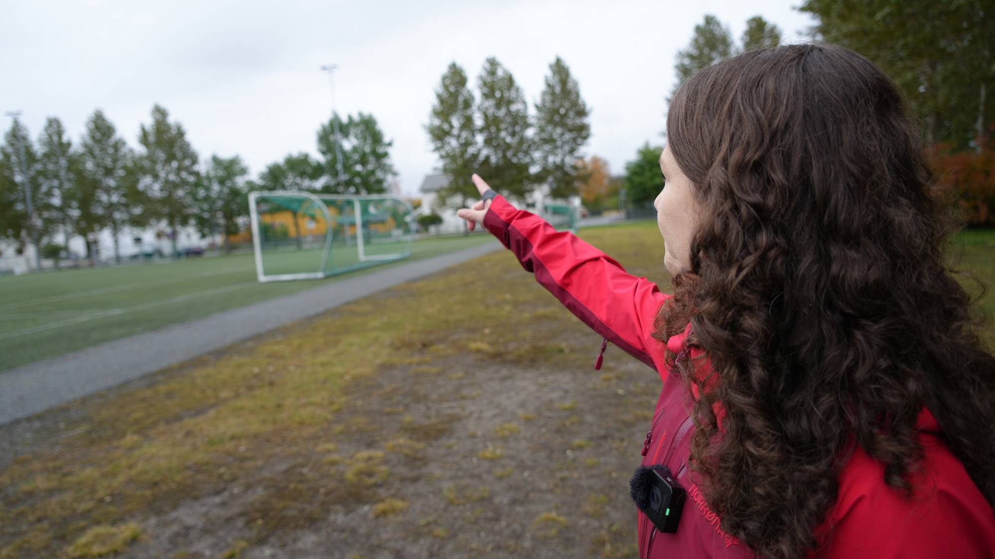 Kvinne i rød jakke peker mot en fotballbane utendørs. (Bildebeskrivelsen er laget av en KI-tjeneste)