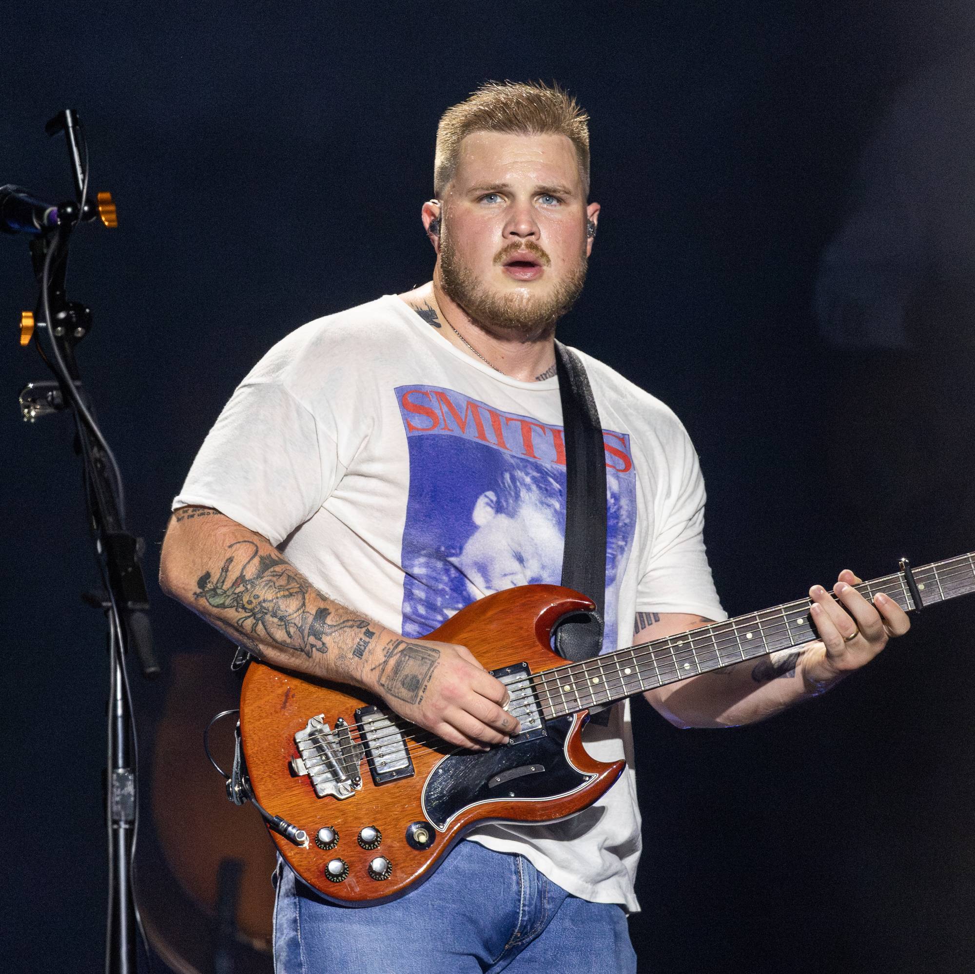 En mann med tatoveringer spiller gitar på en scene. Han har på seg en hvit t-skjorte med trykk og jeans. Mannen er i fokus, mens bakgrunnen er mørk og uskarpt. En mikrofonstativ er synlig til venstre for mannen. (Bildebeskrivelsen er laget av en KI-tjeneste)