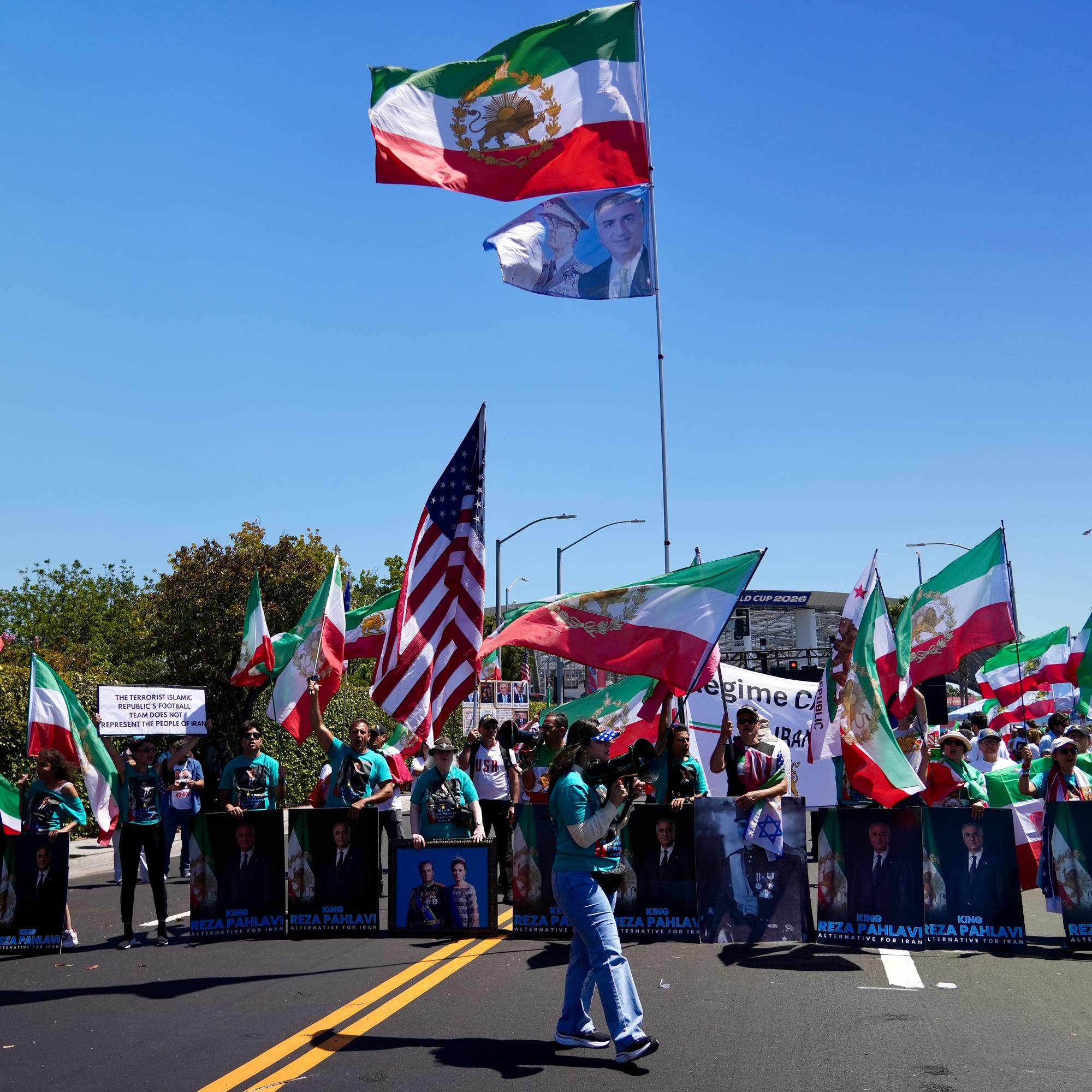 En gruppe mennesker demonstrerer med iranske flagg og bilder av Reza Pahlavi. En kvinne holder et skilt hvor det står "The terrorist Islamic Republic's football team does not represent people of Iran". Flere amerikanske flagg sees i mengden. (Bildebeskrivelsen er laget av en KI-tjeneste)