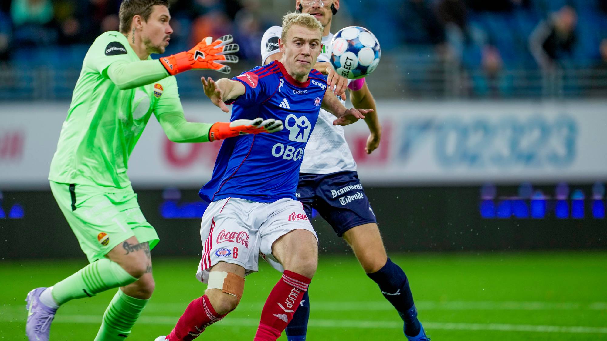 En grønnkledd målvakt forsøker å blokkere en fotball mens en spiller i blått nærmer seg. Spilleren i blått har en bandasje på venstre kne. Bak dem er en spiller i hvitt med mørke bukser. Omgivelsene viser et fotballstadion med tilskuere i bakgrunnen.