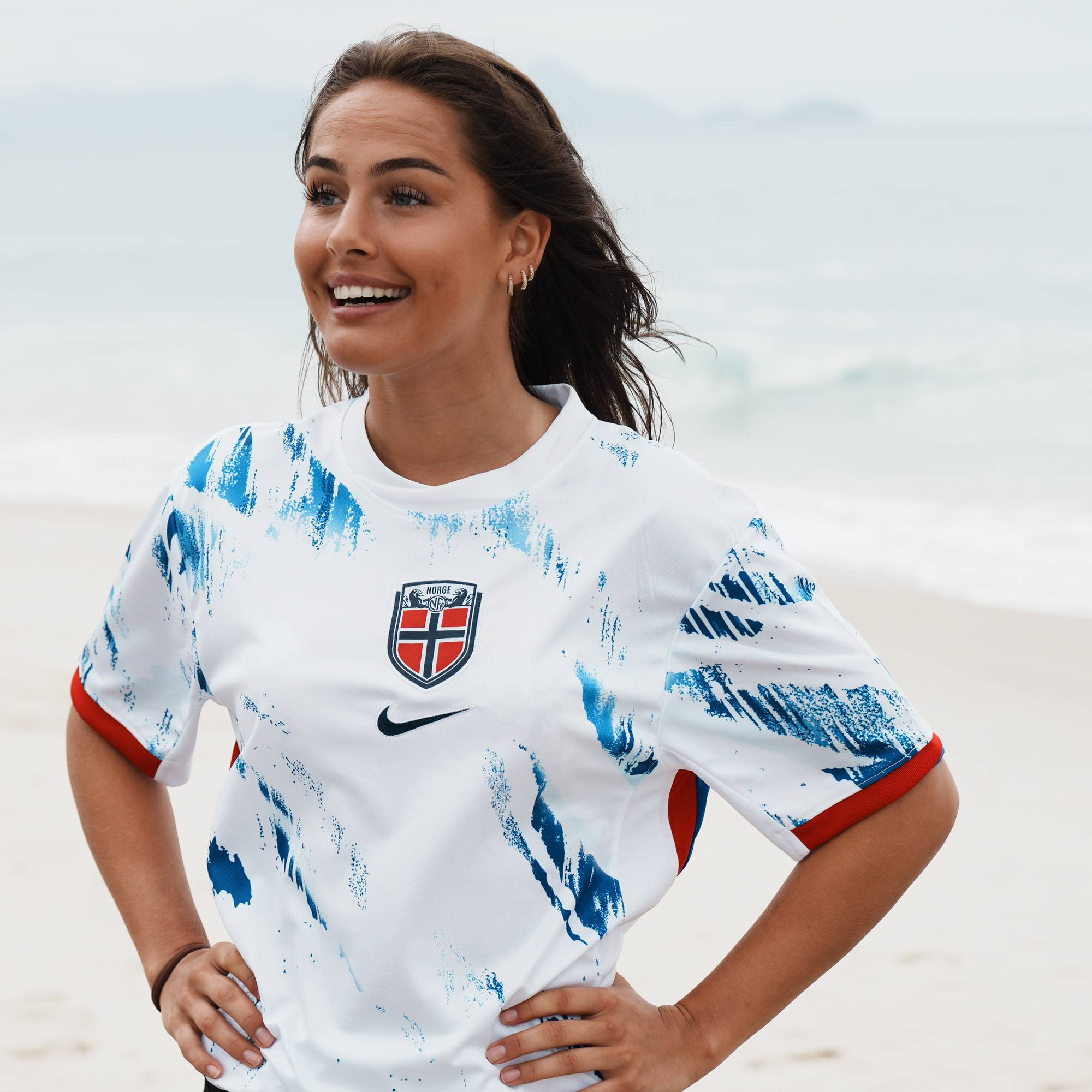 Kvinne i norsk landslagsdrakt for fotball står på en strand og smiler. (Bildebeskrivelsen er laget av en KI-tjeneste)