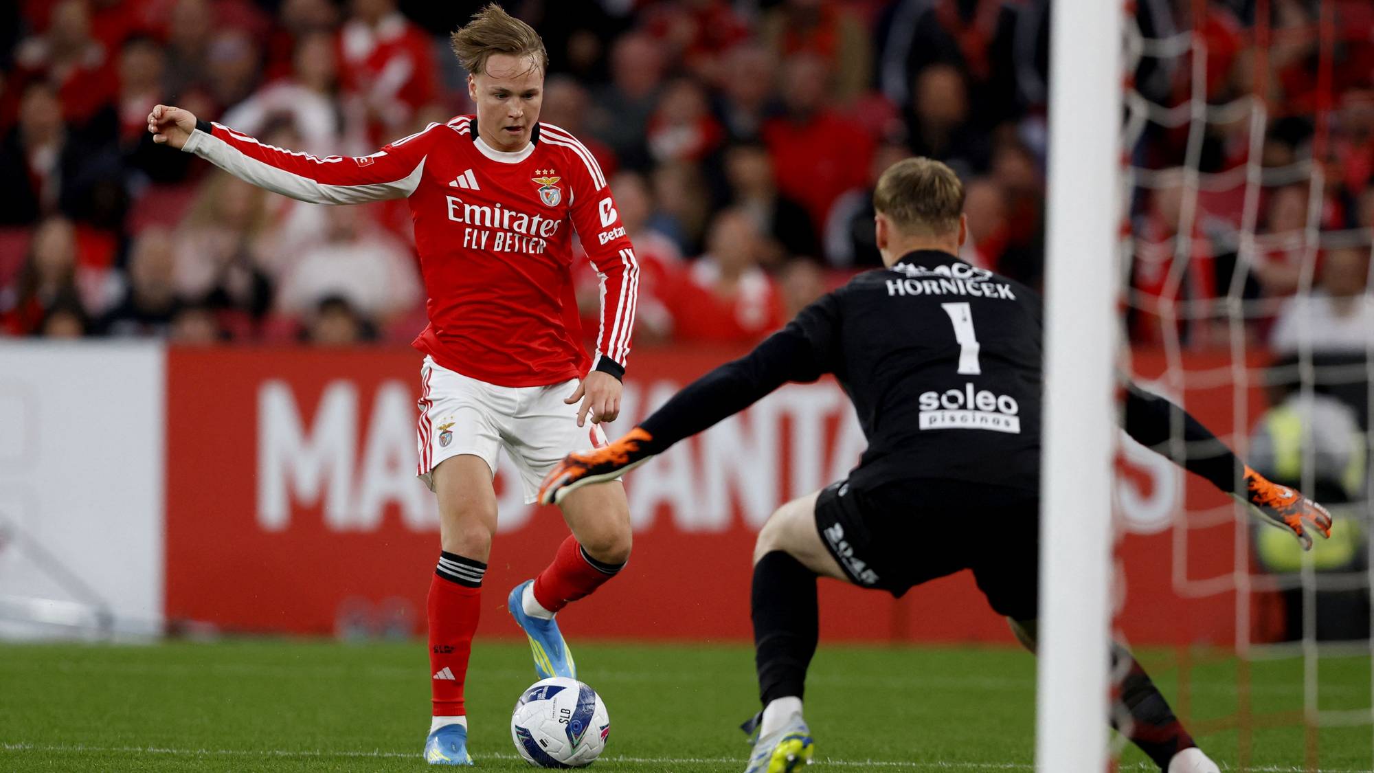 En fotballspiller i rød drakt med nummer 10 mottar ballen på gresset. Han dribler mens en keeper i svart drakt med nummer 1 forsøker å stoppe ham. Tilskuere ses i bakgrunnen. (Bildebeskrivelsen er laget av en KI-tjeneste)