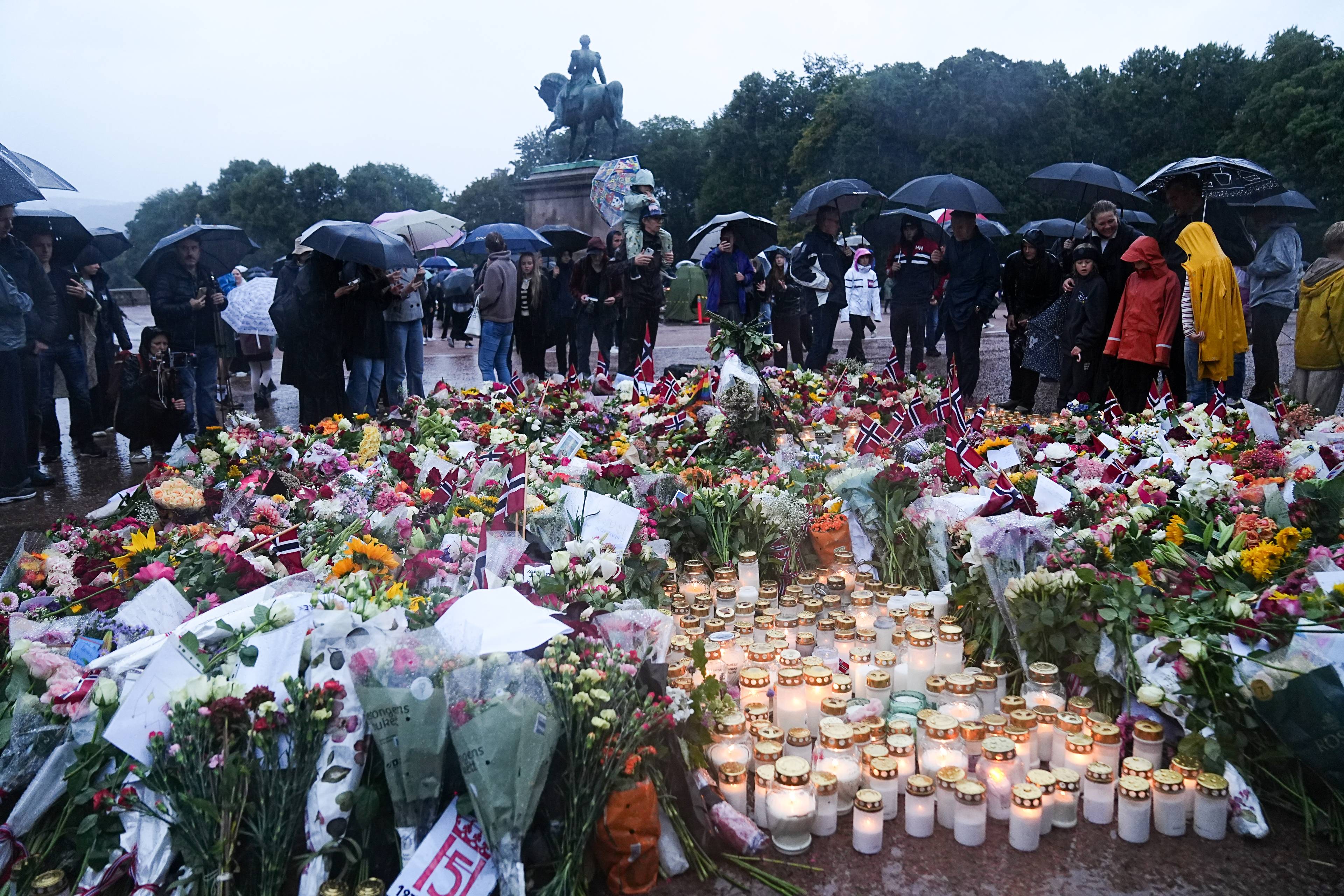 Folk med paraplyer samlet foran blomster og tente lys ved Karl Johan-statuen i Oslo. (Bildebeskrivelsen er laget av en KI-tjeneste)