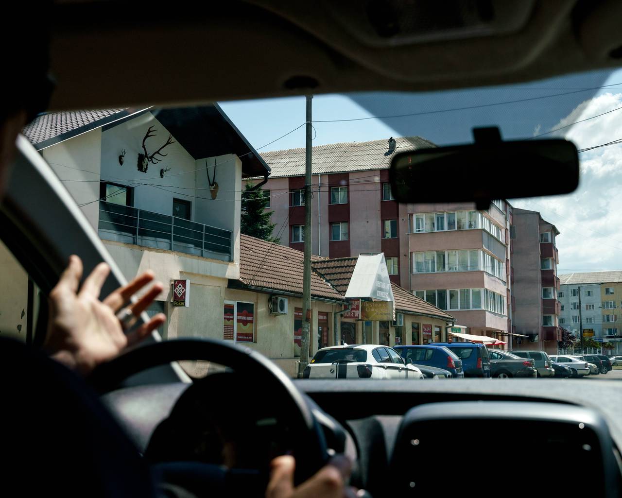 A woman drives a car along a road in Romania.  Through the windshield you can see a vegetable shop and a Chinese restaurant.