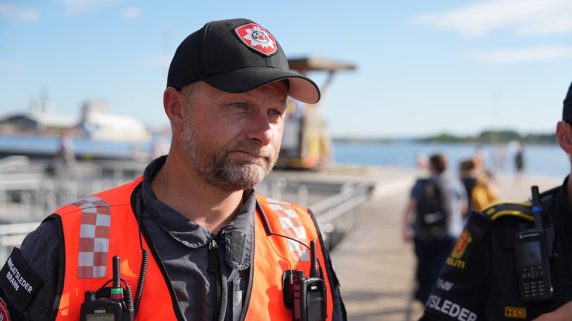 Innsatsleder fra brannvesenet og en politibetjent ved vannkanten på en solskinnsdag. (Bildebeskrivelsen er laget av en KI-tjeneste)