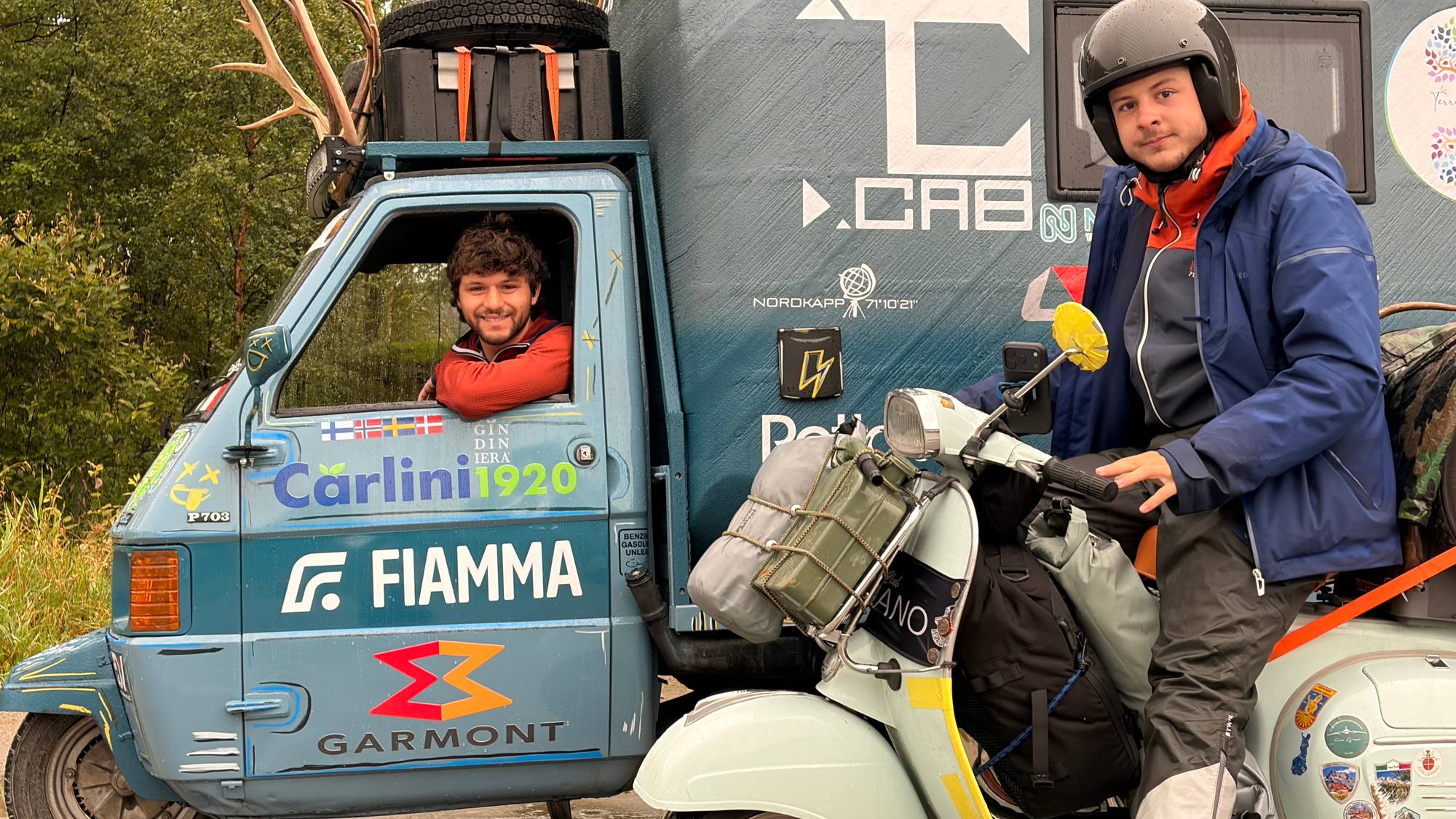 De to vennene Chizzo Pietroo og Furlani Francesco kjører hver sin italienske moped. En tradiosjonell og en mopedvarebil omgjort til en minibobil. Begge sitter på kjøretøyene og smiler til baker.