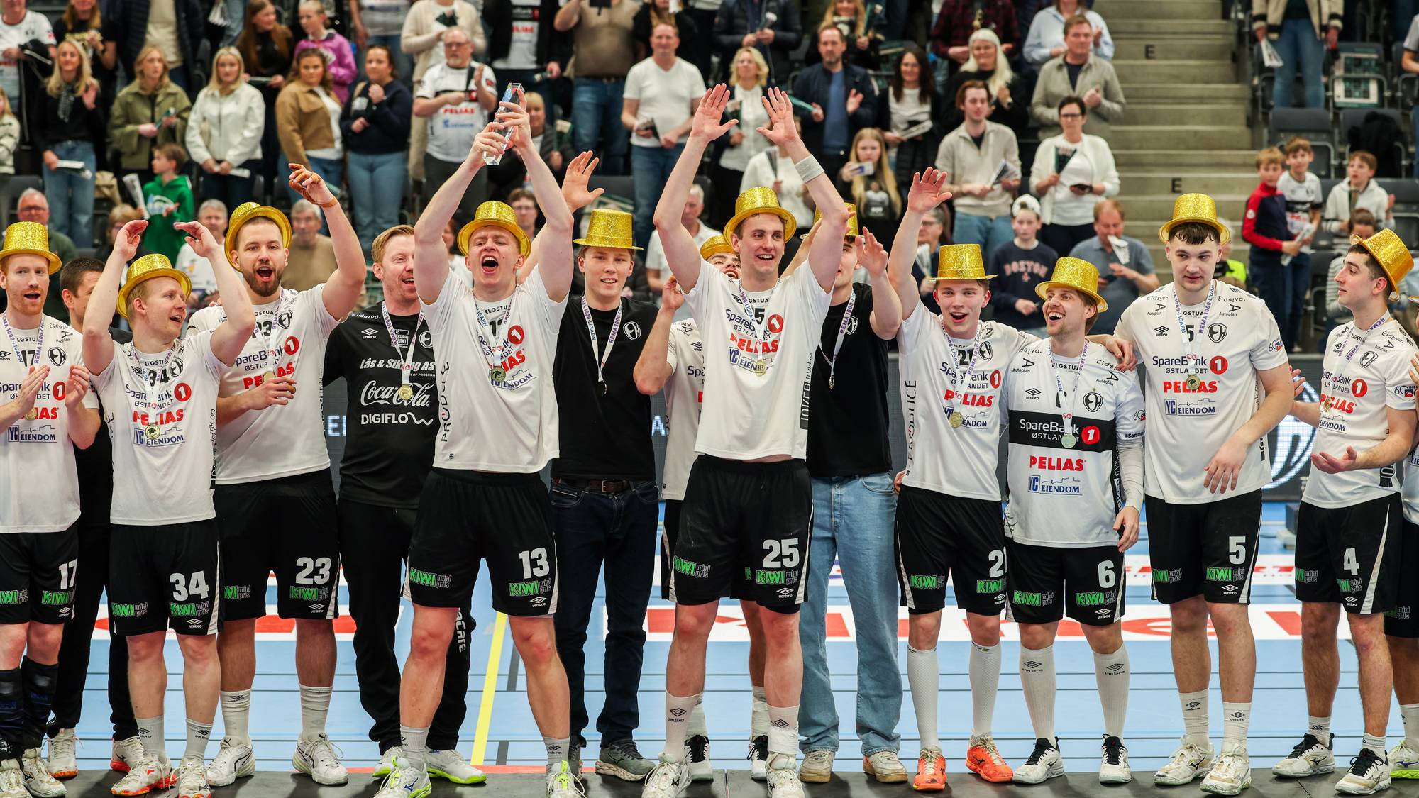 En gruppe idrettsutøvere med gullhatter feirer på et podium. Flere av dem holder opp flasker og løfter armene i været. Publikum er synlig i bakgrunnen på tribunen. (Bildebeskrivelsen er laget av en KI-tjeneste)