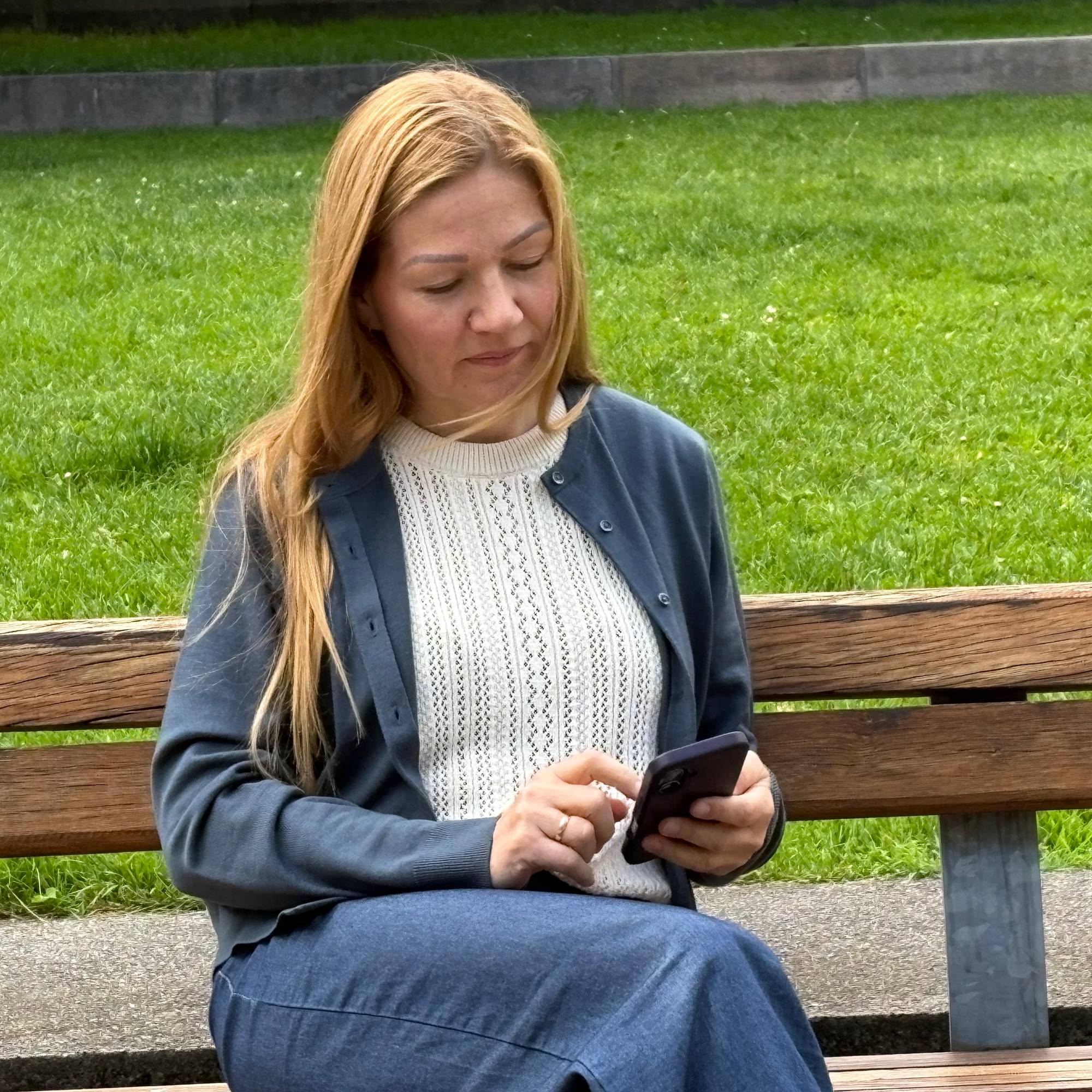 Kvinne med langt, brunt hår sitter på en benk i en park og ser på telefonen sin.