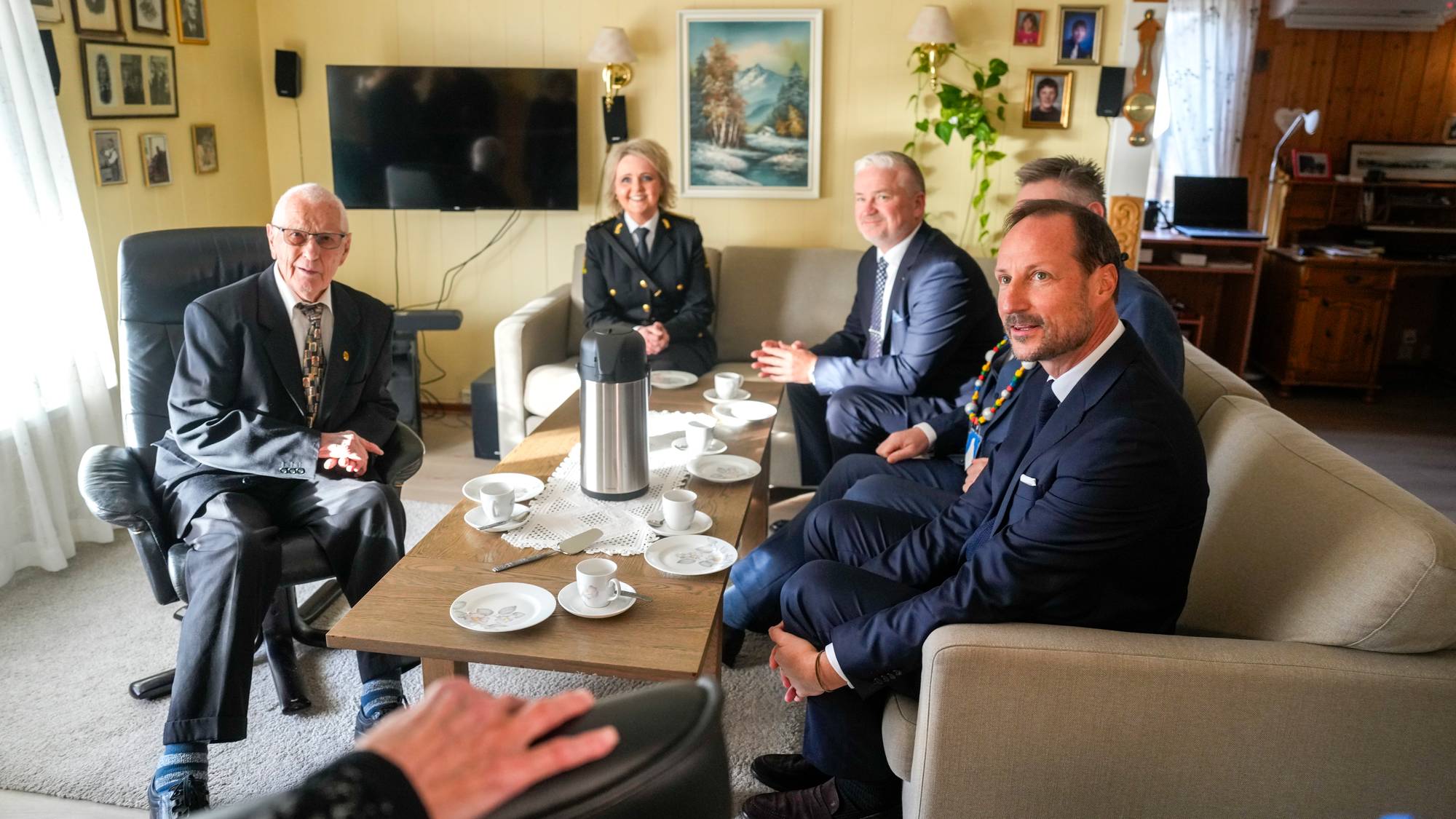 Kronprins Haakon sitter i en samtale med tre andre personer i en stue. (Bildebeskrivelsen er laget av en KI-tjeneste)