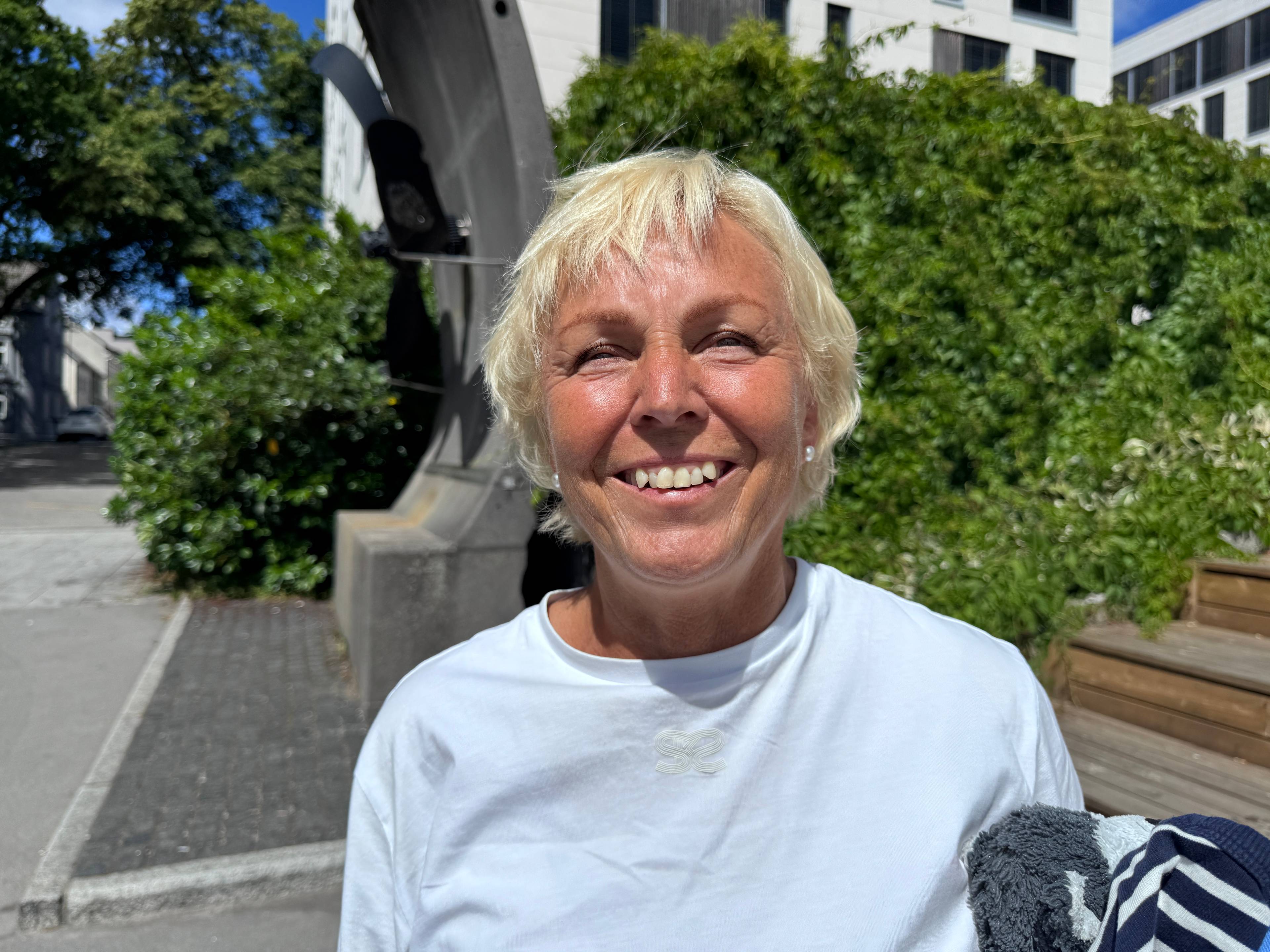 En kvinne med lyst, kort hår og hvit t-skjorte smiler. I bakgrunnen ser man en vei og grønne busker.
