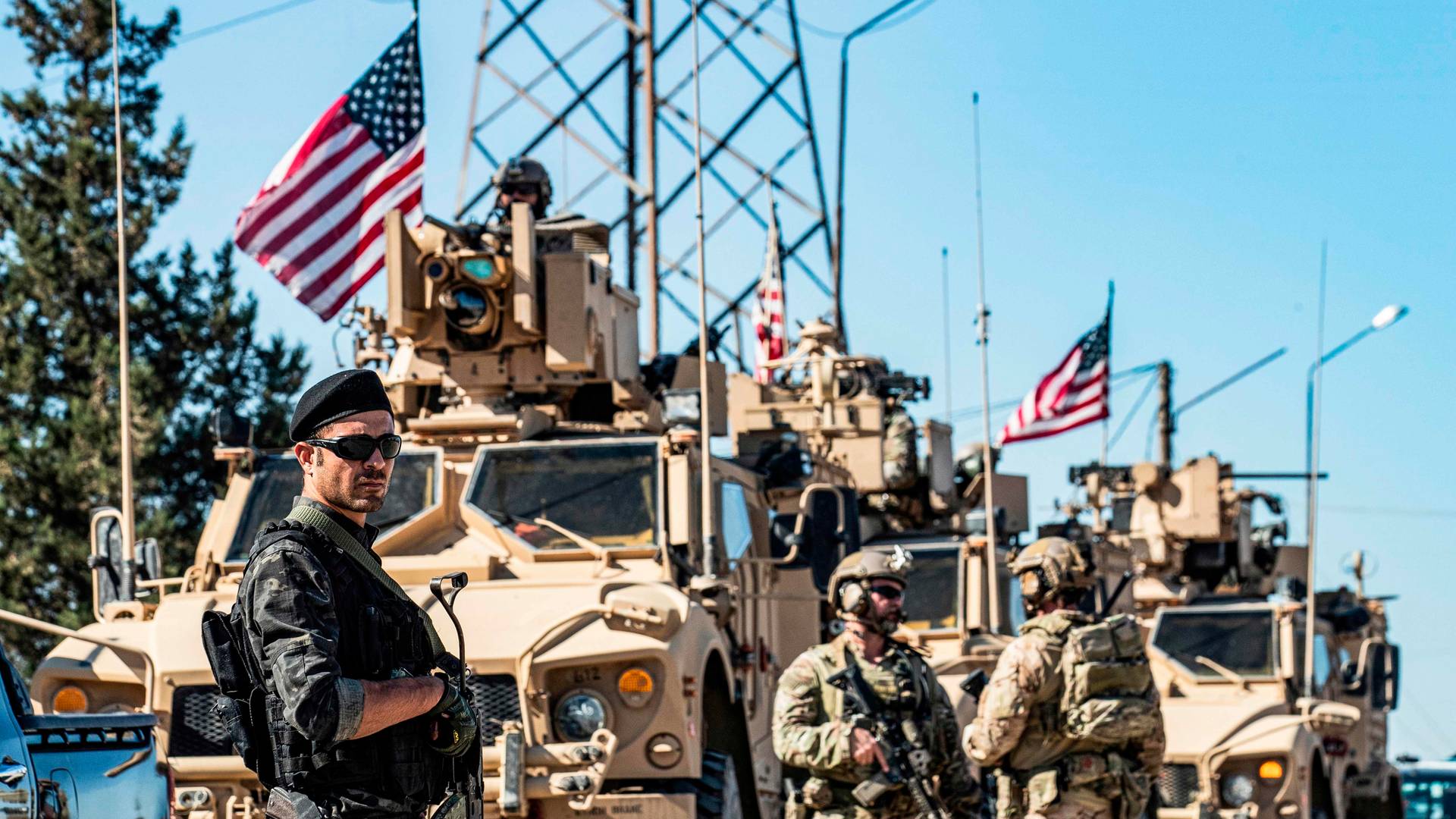 A⁤ member (left) ‍of the Kurdish People's Protection Units (YPG) stands guard in‌ front of US armored vehicles and soldiers on ​patrol near an oil well in Rumaylan ⁤(Rmeilan)⁣ in northeastern Syria's Hasakeh province on ⁤November‍ 6, 2019.