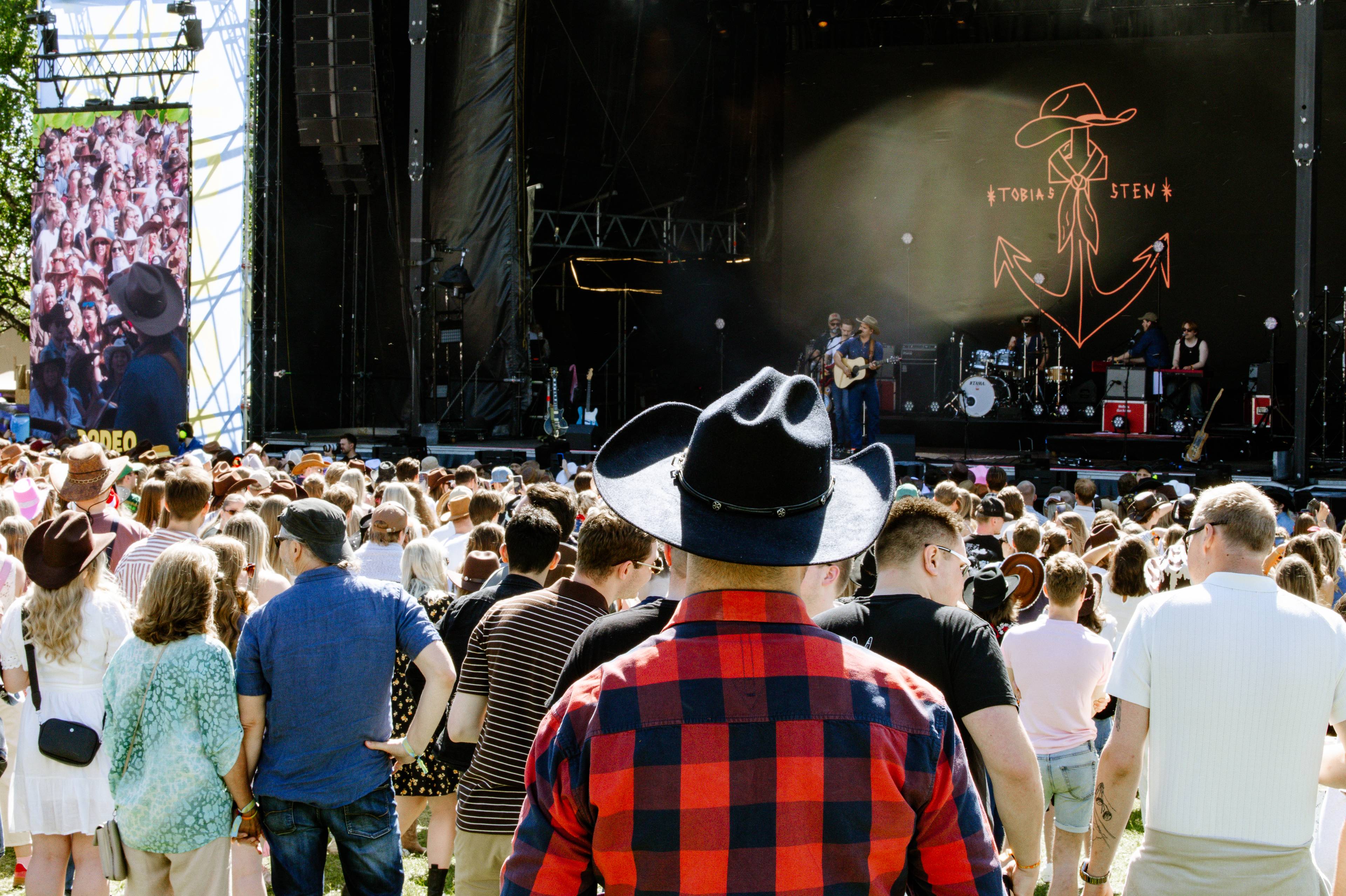 Festivalpublikum ser på en konsert. Et band spiller på en scene med en stor lysende logo av et anker og en cowboyhatt. Til venstre for scenen er det en storskjerm med bilder av mennesker. Mange i publikum har på seg cowboyhatter. (Bildebeskrivelsen er laget av en KI-tjeneste)