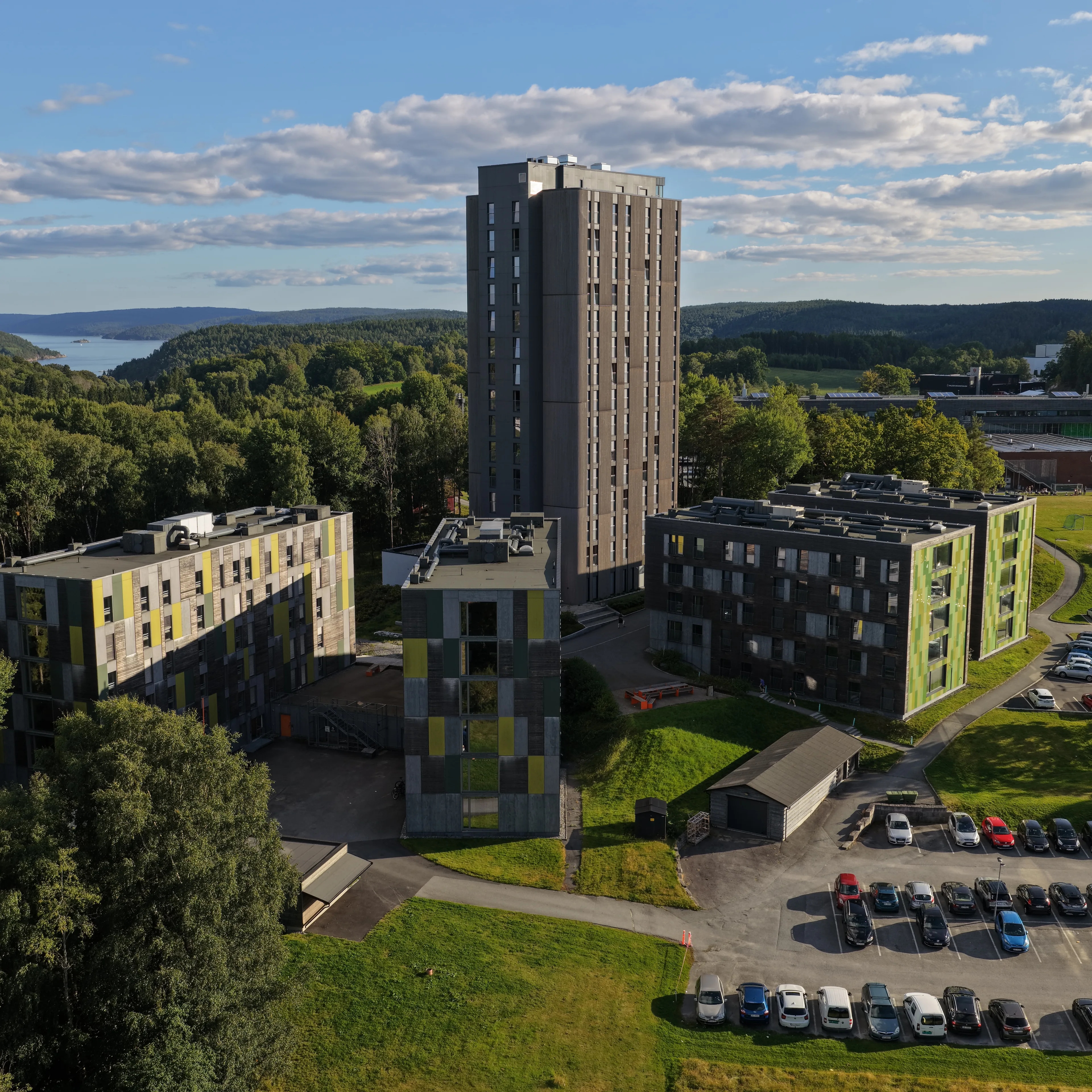 Flyfoto av et høyt grått boligtårn og tilhørende leilighetsbygg omgitt av skog, plen og parkeringsplass. (Bildebeskrivelsen er laget av en KI-tjeneste)
