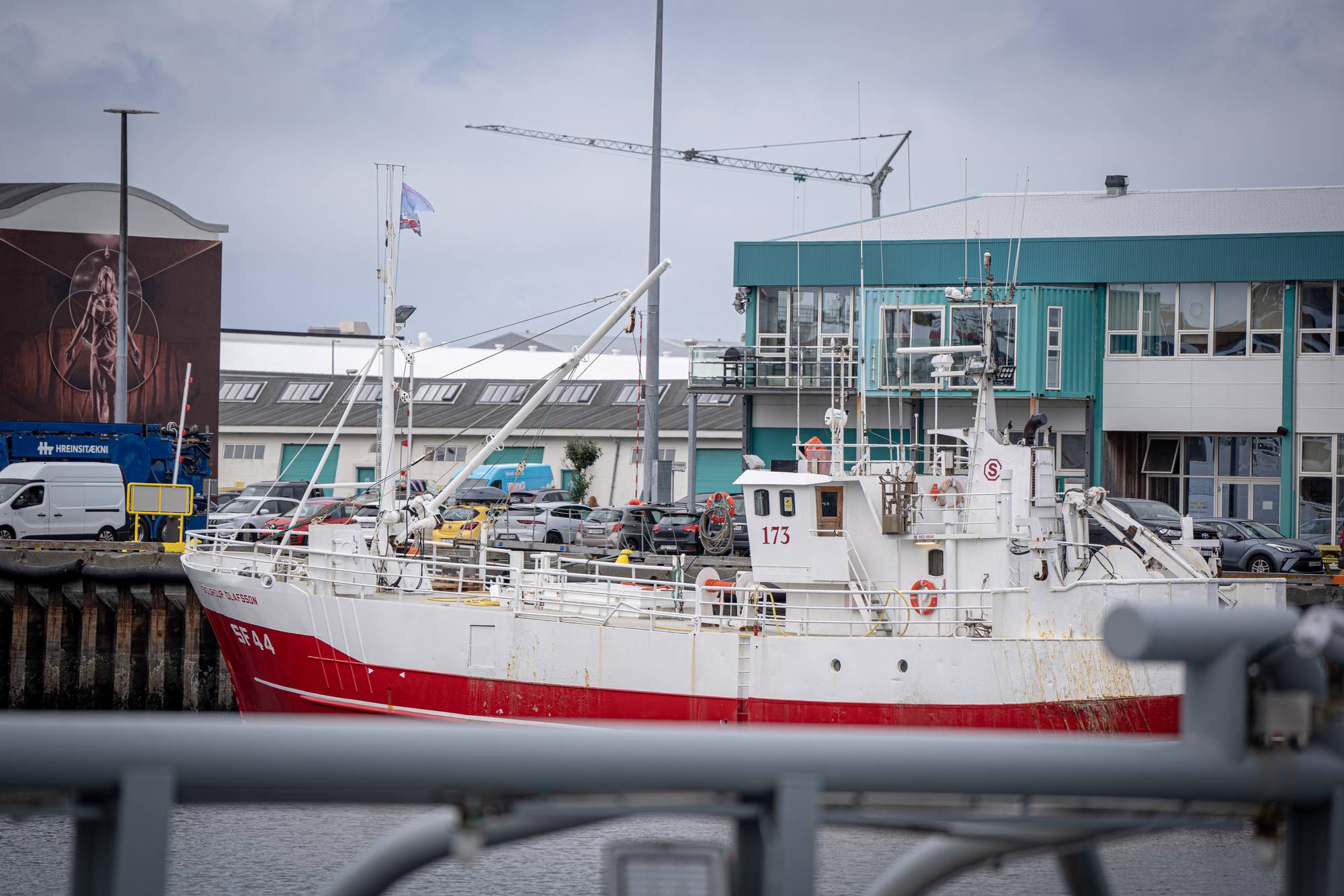 Hvit og rød fiskebåt ligger til kai ved en bygning i en havn. (Bildebeskrivelsen er laget av en KI-tjeneste)